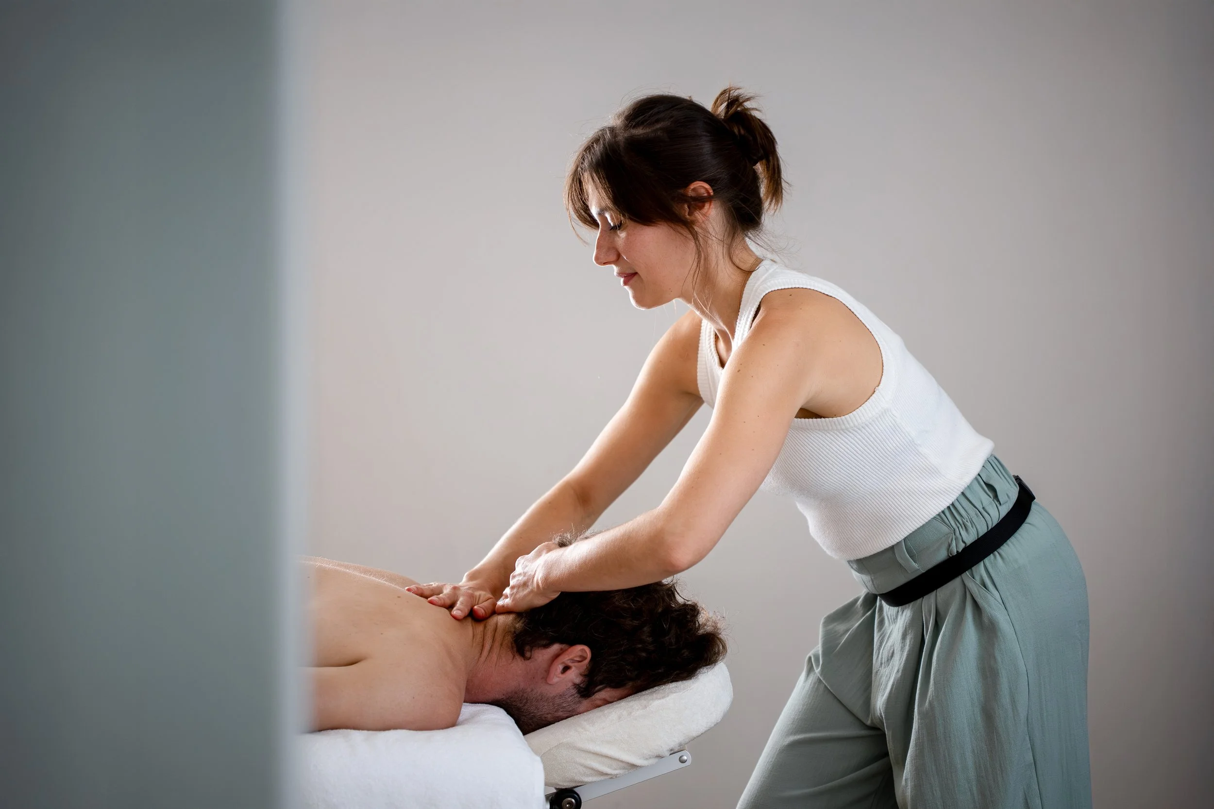 Une femme donne un massage de la nuque à un homme allongé sur une table de massage, dans un environnement calme et simple. Lissages, mains alternées, remontant le long du haut du dos et de la nuque. 
