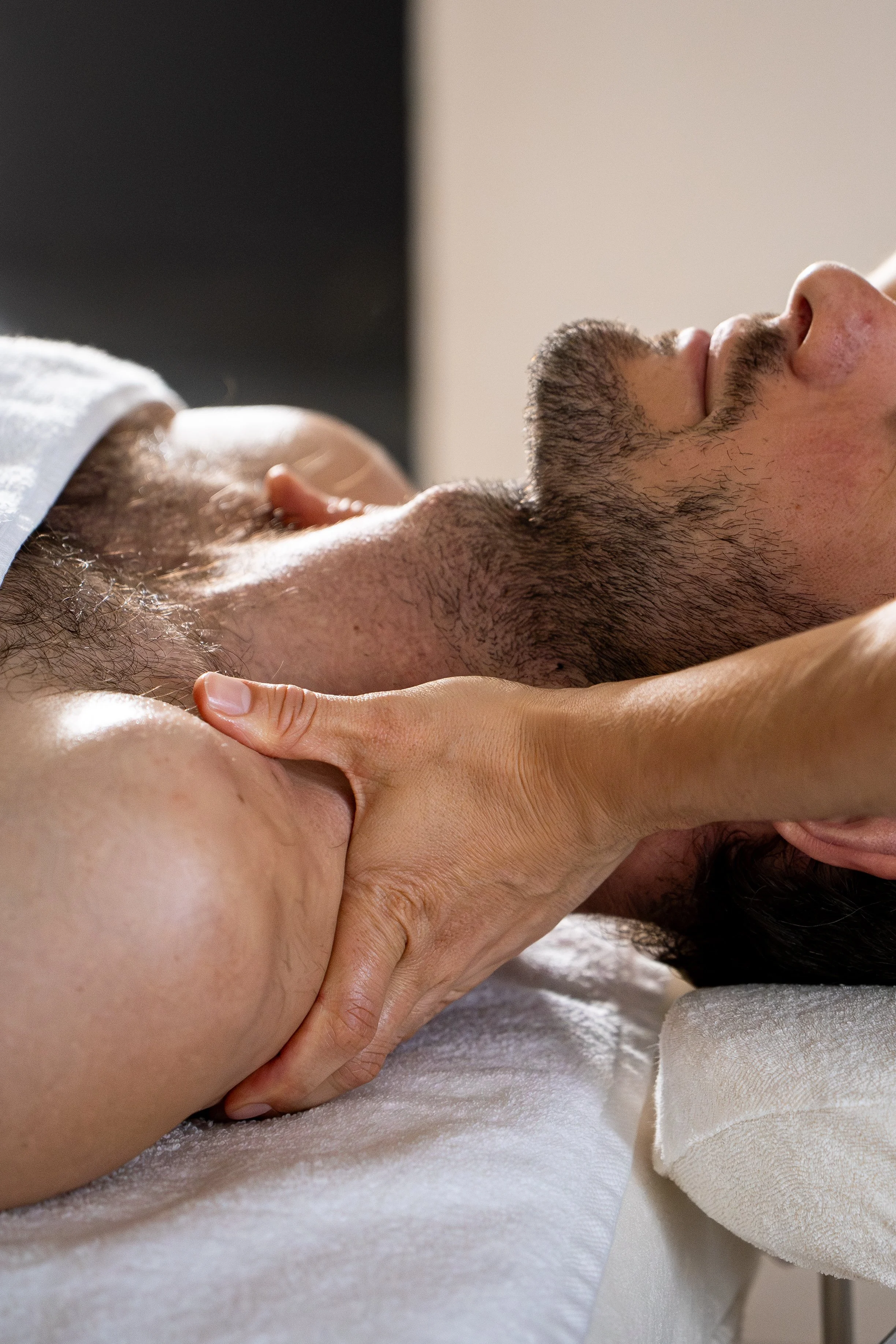Un homme allongé sur une table de massage avec un drap blanc reçoit un massage du cou, des trapèzes, des épaules.  La personne qui donne le massage utilise ses mains et de l'huile de massage.