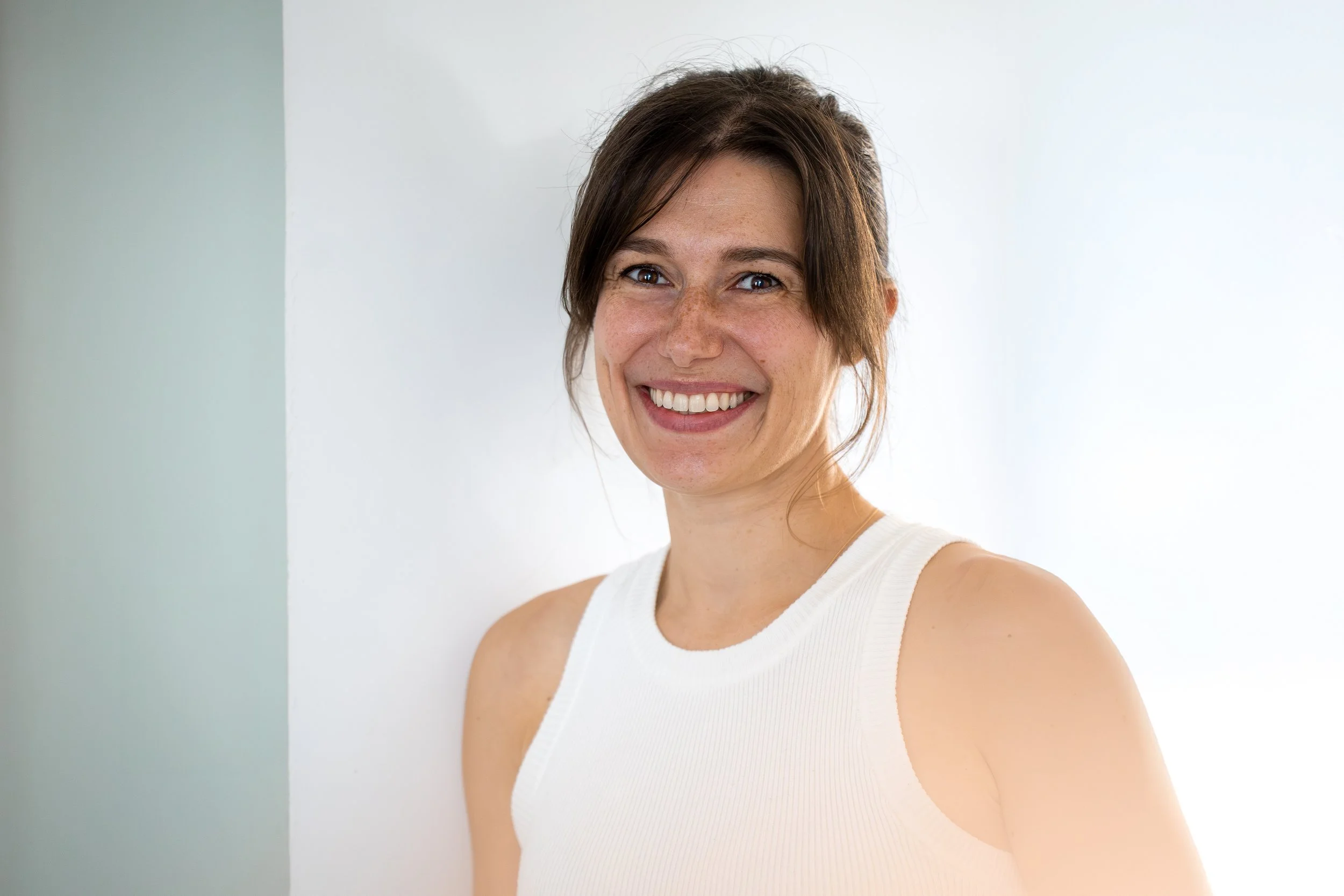 Une femme souriante avec des cheveux bruns, portant un haut blanc, posant contre un mur blanc.