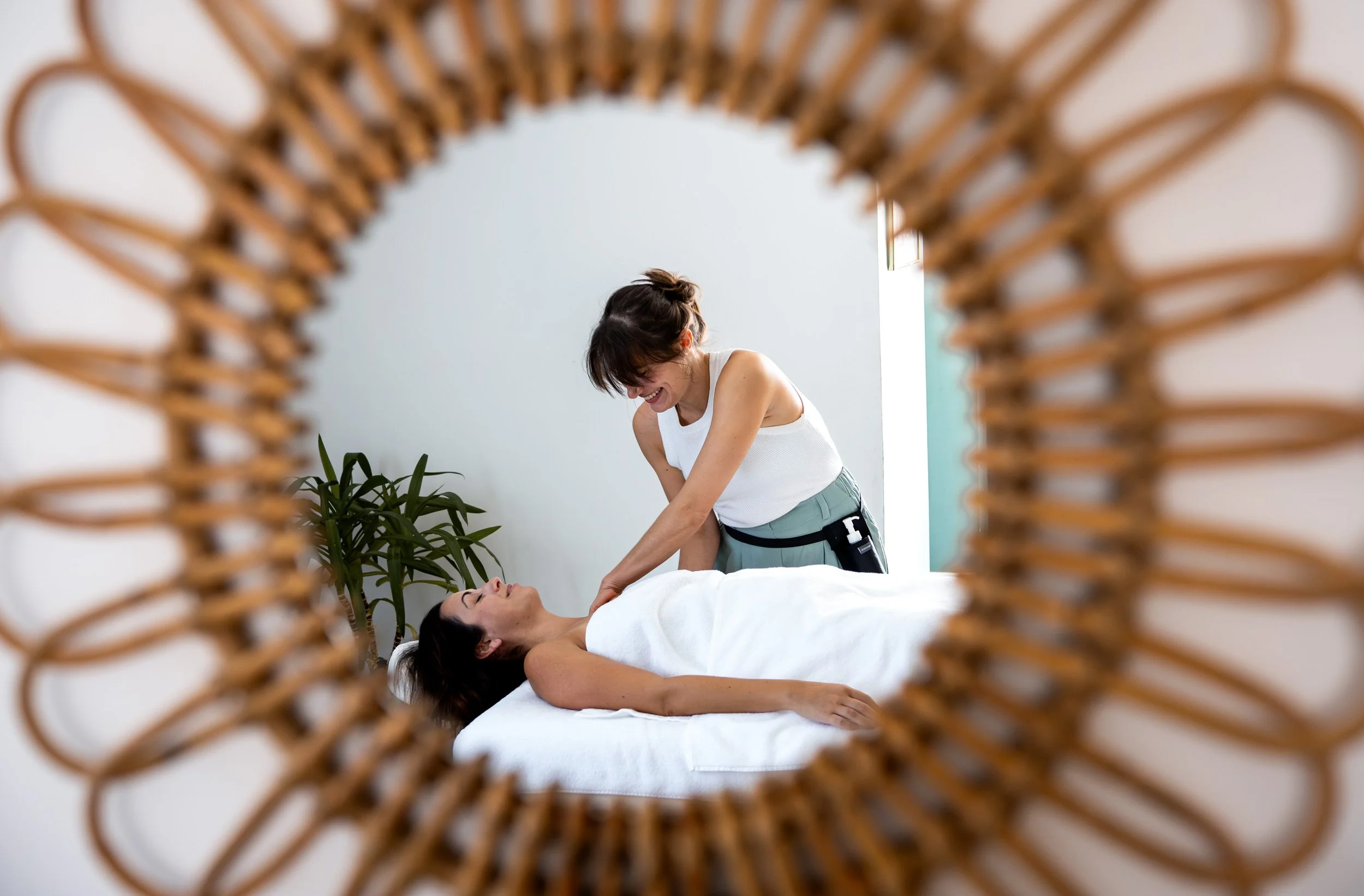 Une femme, allongée sur le dos sur une table de massage, recevant un massage de l'épaule et du bras, vue à travers un miroir en rotin.