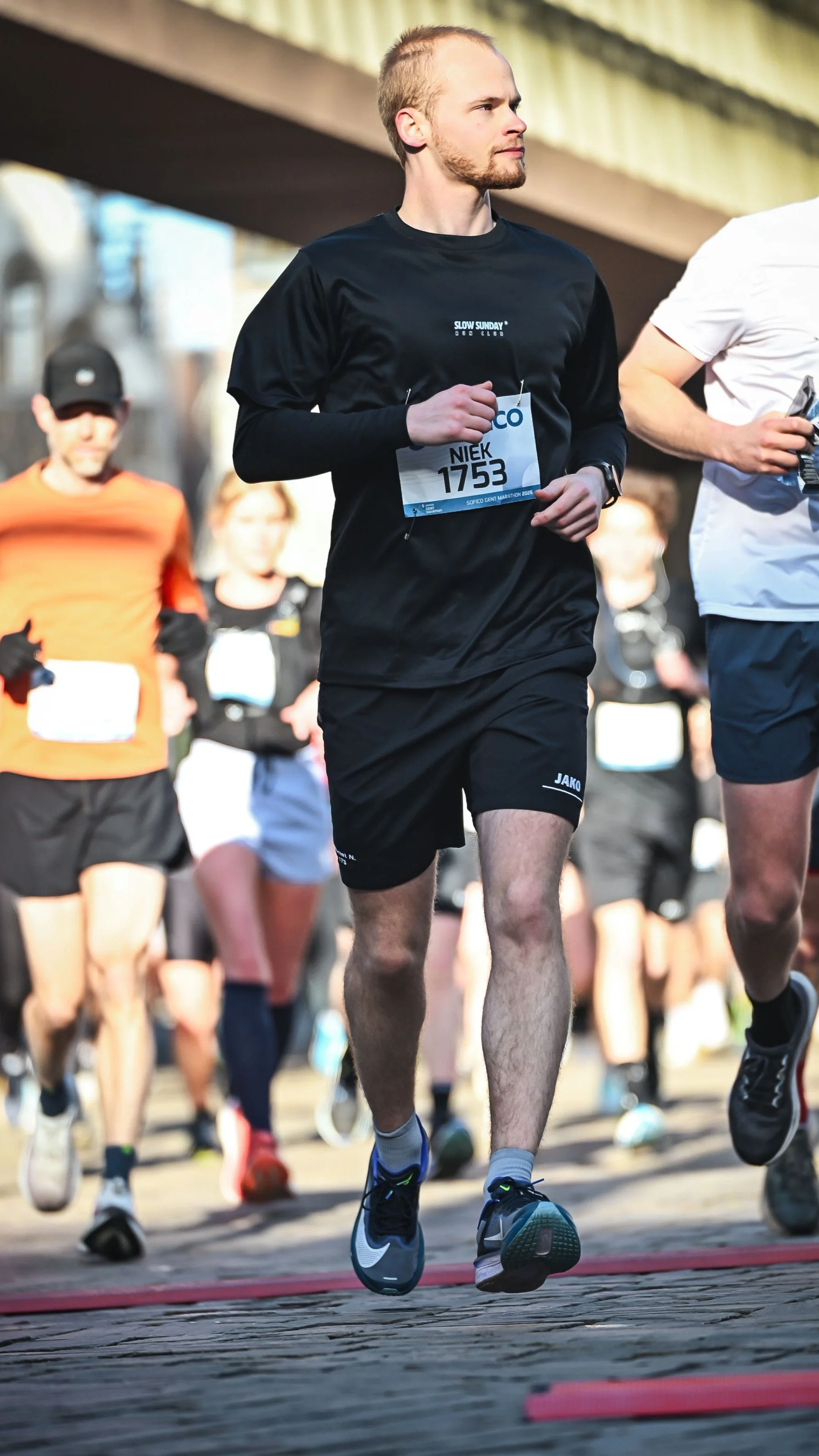 Man in black long-sleeve shirt and shorts participating in a marathon, running with other runners on a city street.
