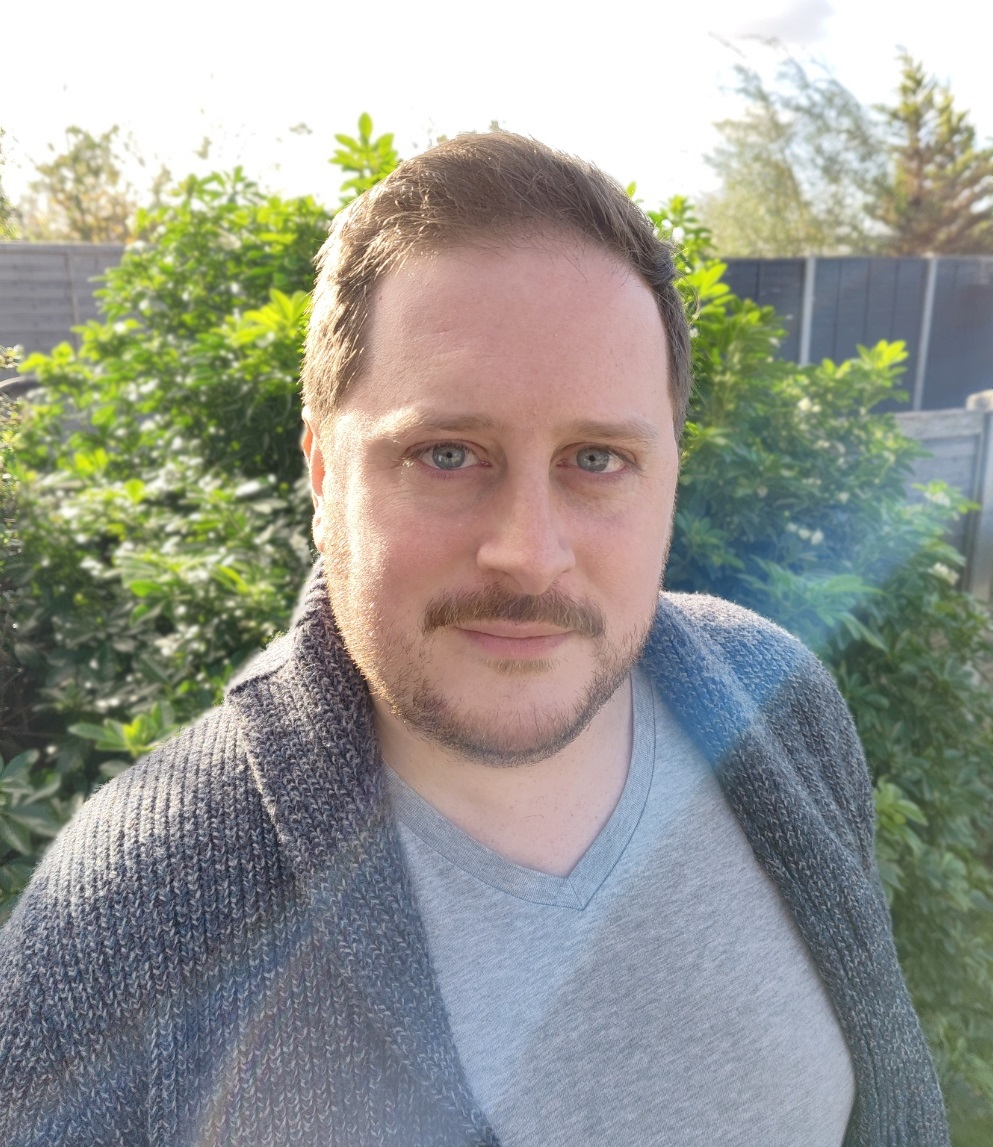 A man with light skin, blue eyes, and brown hair takes a selfie outdoors during daytime. He is wearing a gray shirt and a dark knitted cardigan. The background features green bushes, a wooden fence, and trees under a partly cloudy sky.