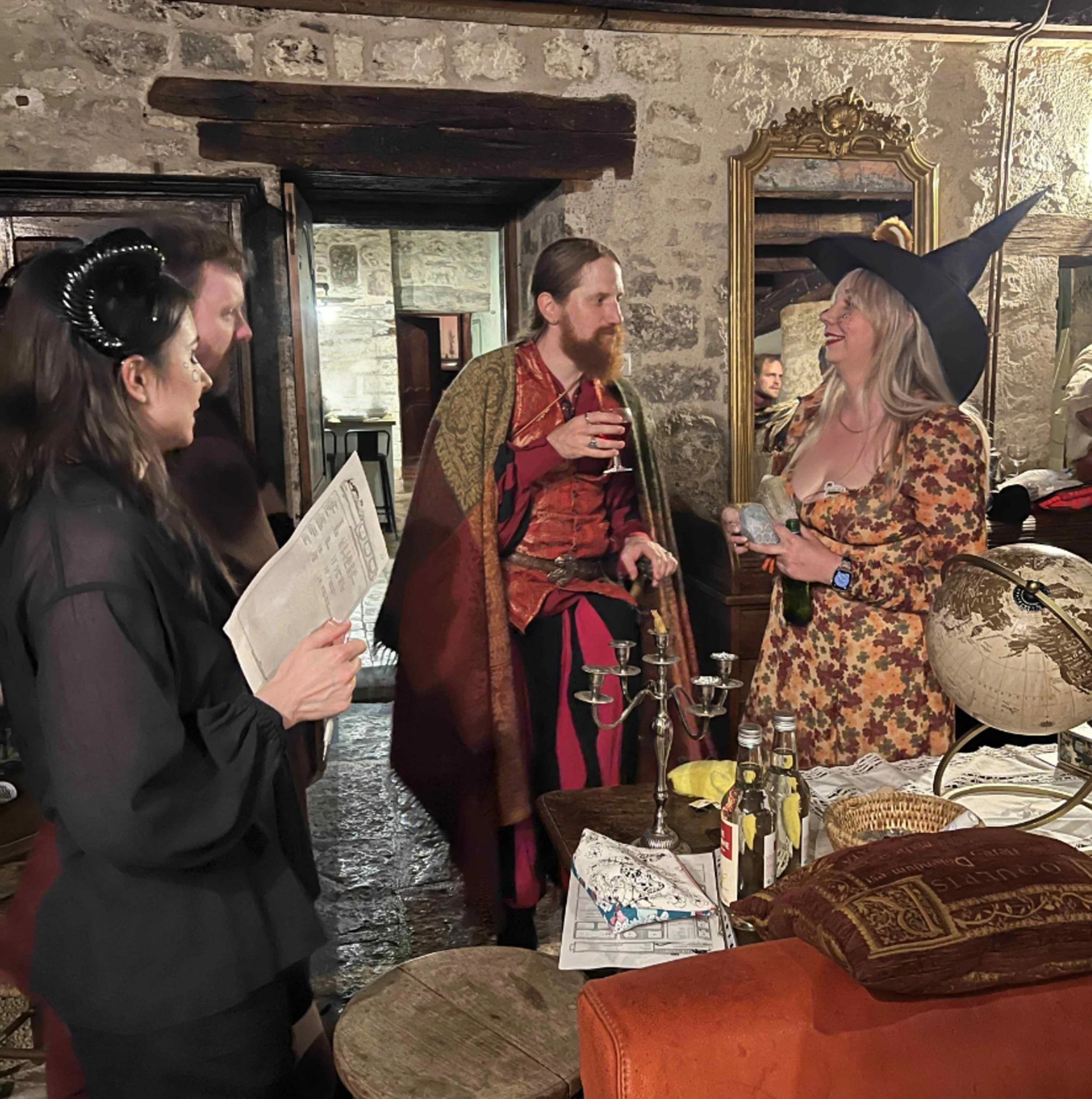 People dressed in costumes at a themed gathering, with a woman in a witch hat and floral dress, and a man in medieval-style clothing, engaged in conversation in a rustic, stone-walled room.