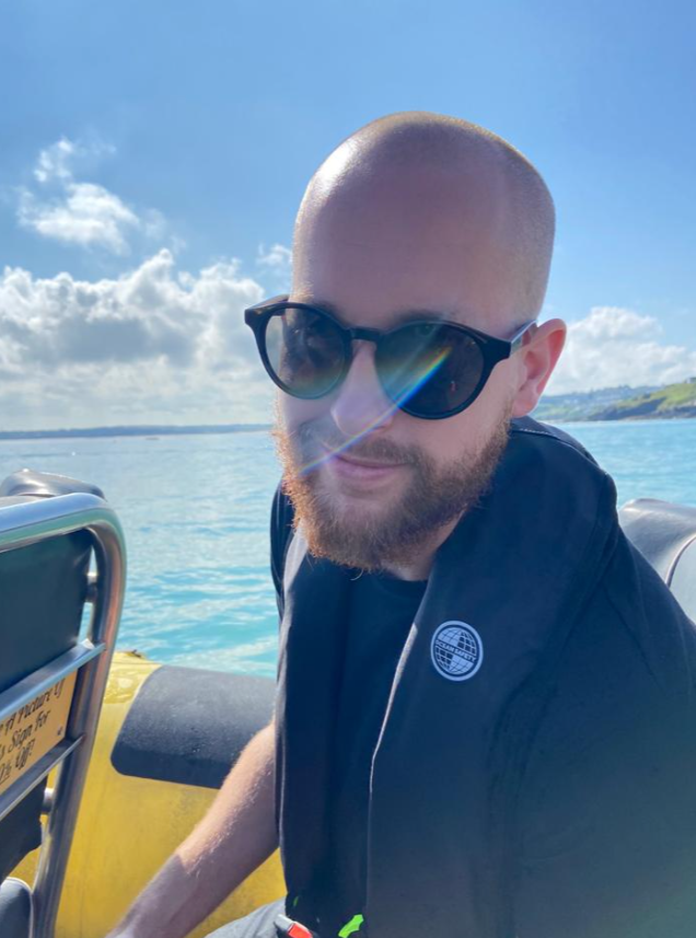 A man with a shaved head and beard wearing sunglasses, sitting outdoors on a boat with water and a blue sky with clouds in the background.