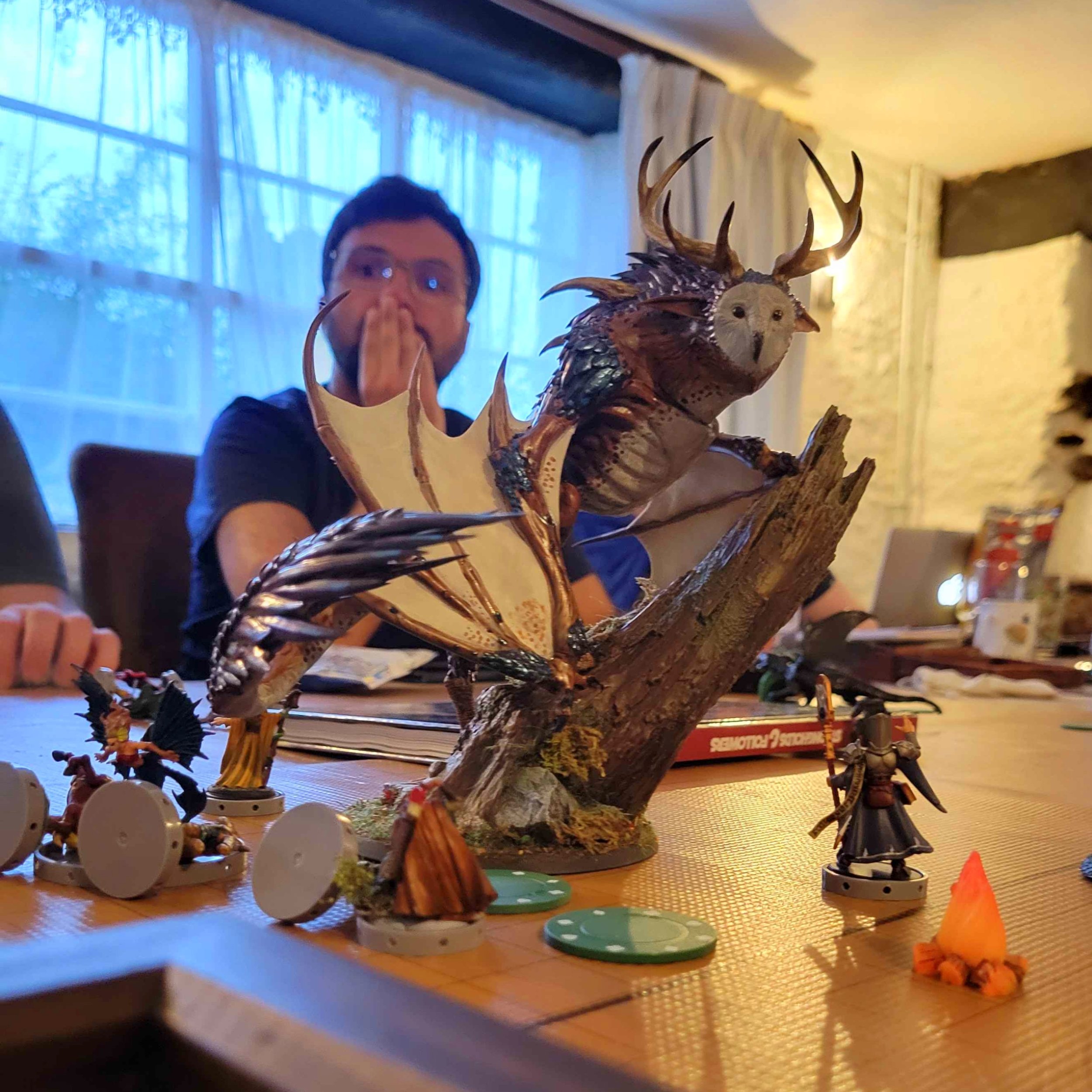 A person seated at a table with tabletop gaming miniatures, including a large, detailed owl-like creature with antlers and wings, and a smaller armored figure, along with other game pieces. The person is in the background, with a surprised or excited expression.