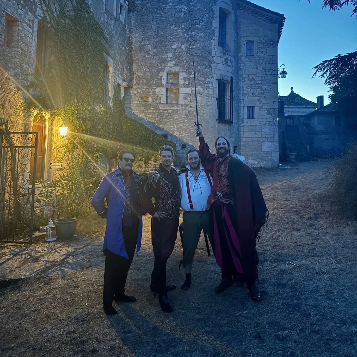Four men dressed in medieval or renaissance costumes standing outside a stone building at dusk, with one holding a sword raised in the air.