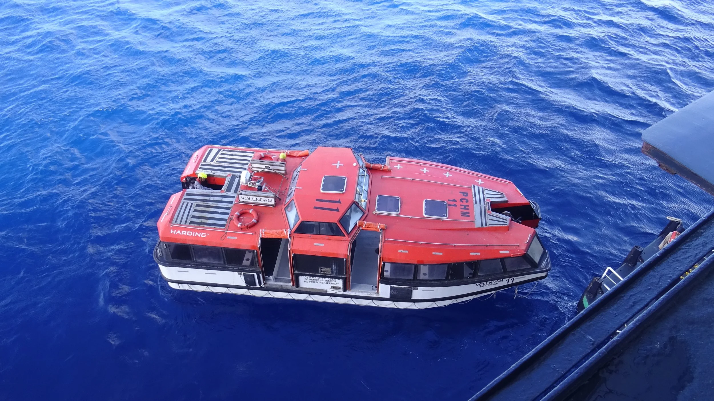 A orange/red lifeboat off the edge of a cruise ship with safety equipment and personnel onboard, floating on blue ocean water, seen from above.