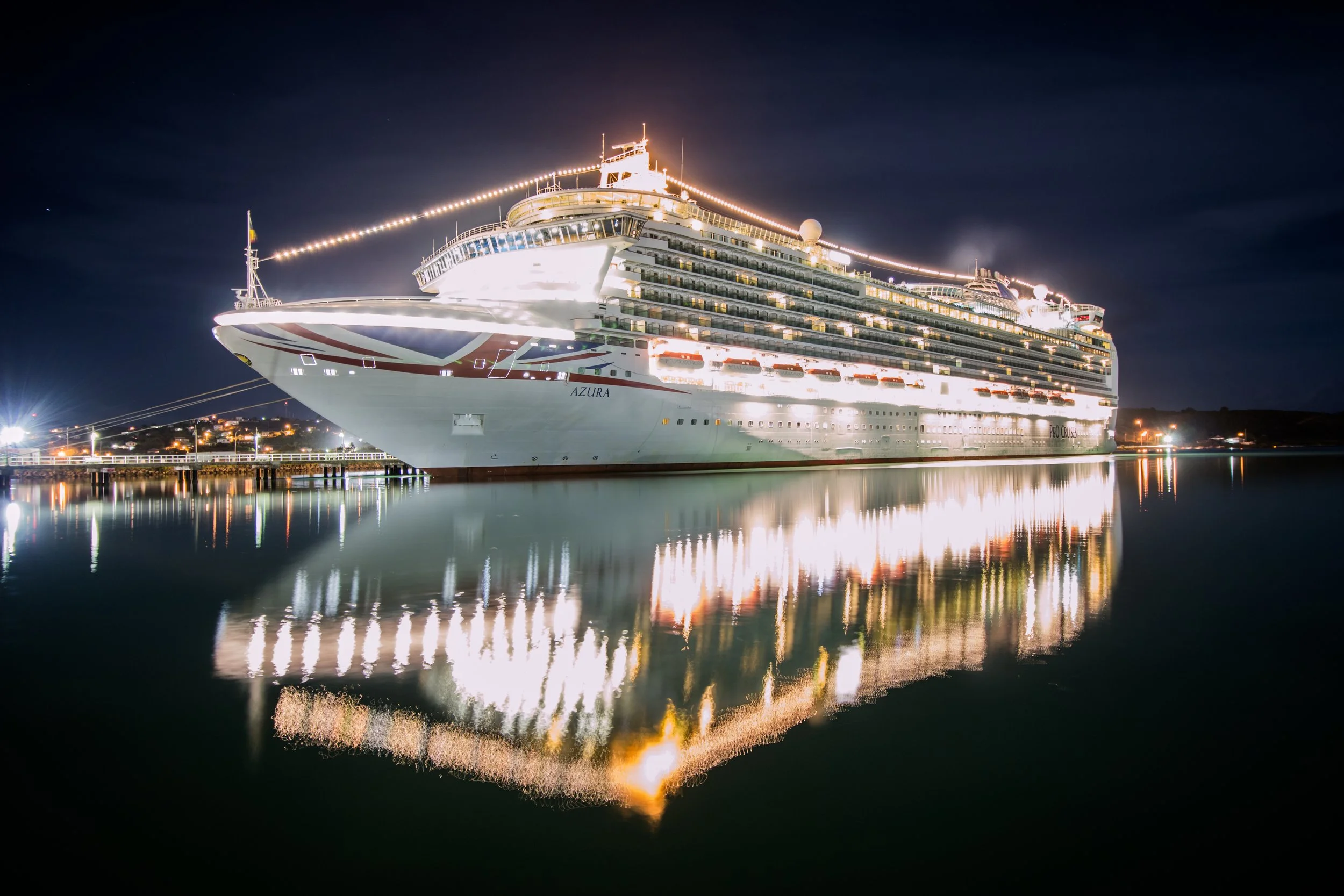 A large cruise ship named Azura docked at night, adorned with bright lights, with its reflection visible on the calm water.