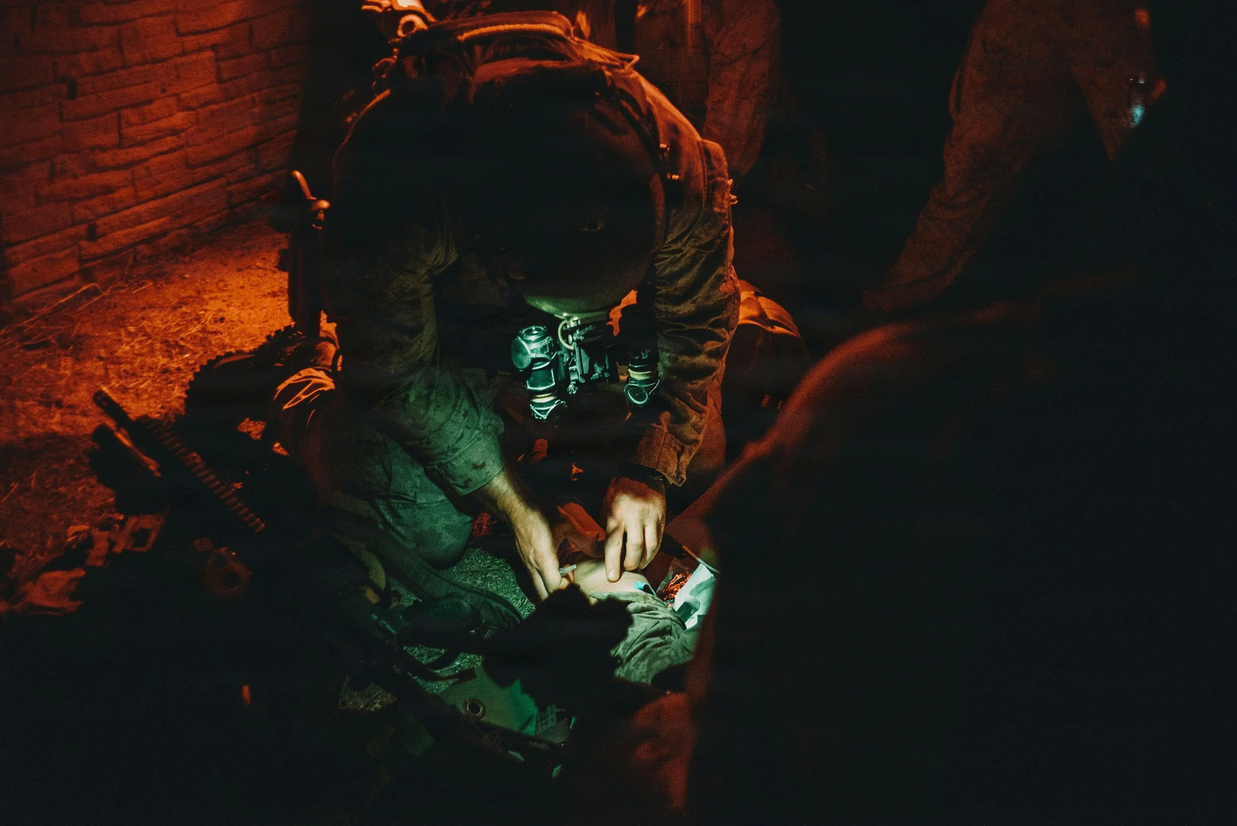 Emergency medical personnel providing aid to a person lying on the ground in a dark setting with red lighting, possibly at night.