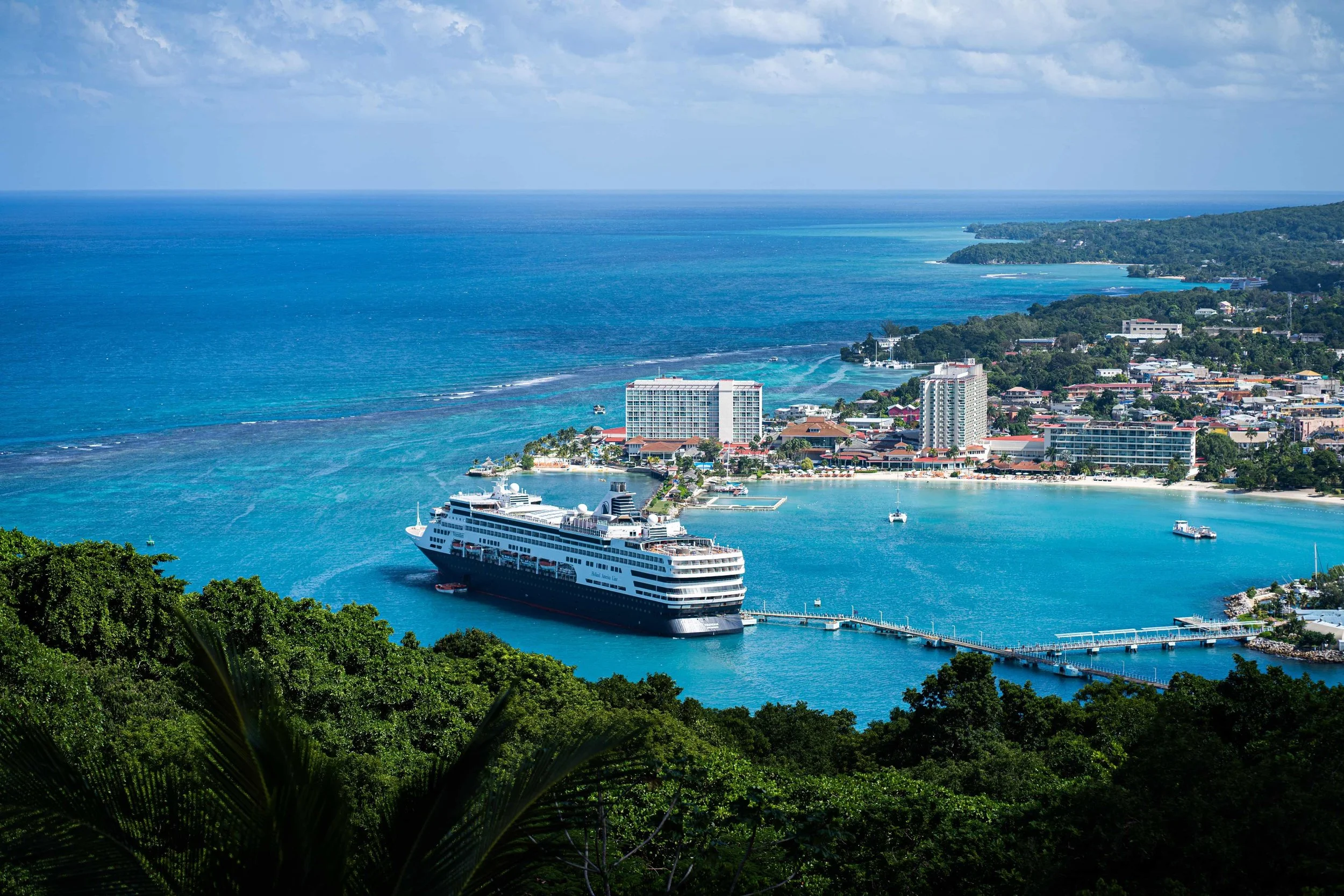 Aerial view of a coastal city in Jamaica, with a large cruise ship docked in the harbor, surrounded by blue ocean waters, green hills, and city buildings.
