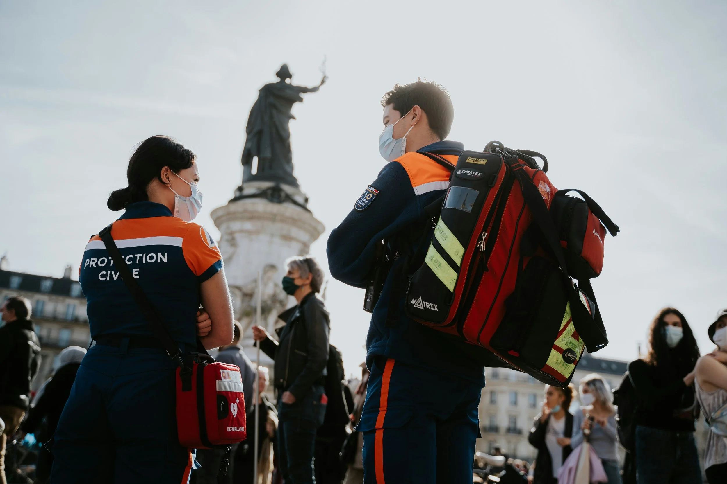 Two emergency responders, a woman and a man, wearing protective masks and uniforms, standing outdoors among a crowd, with a statue in the background.