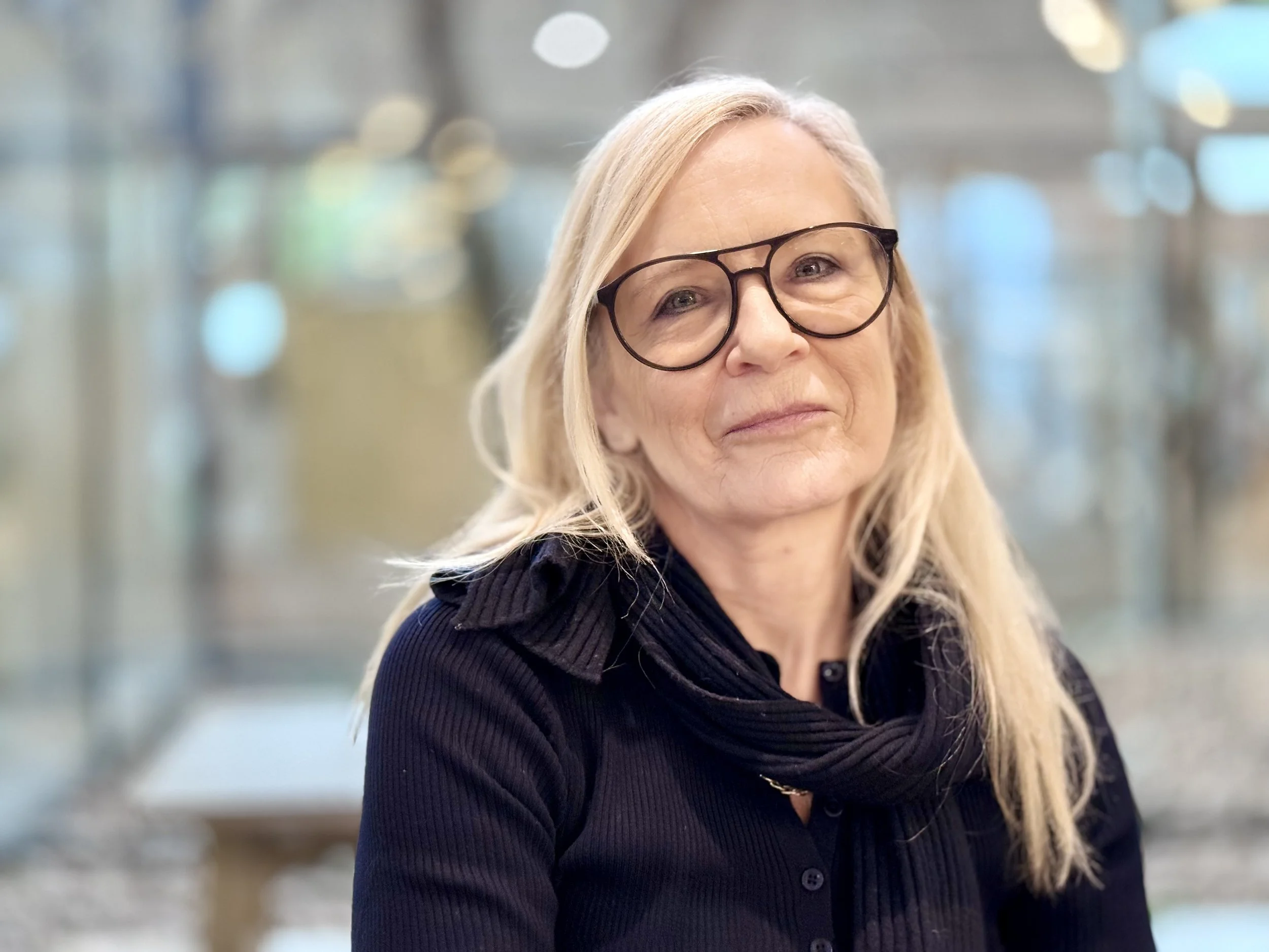 Eine reife Frau mit langen blonden Haaren und Brille, die einen dunklen Pullover und einen Schal trägt, steht in einem Innenraum vor unscharfem Hintergrund.