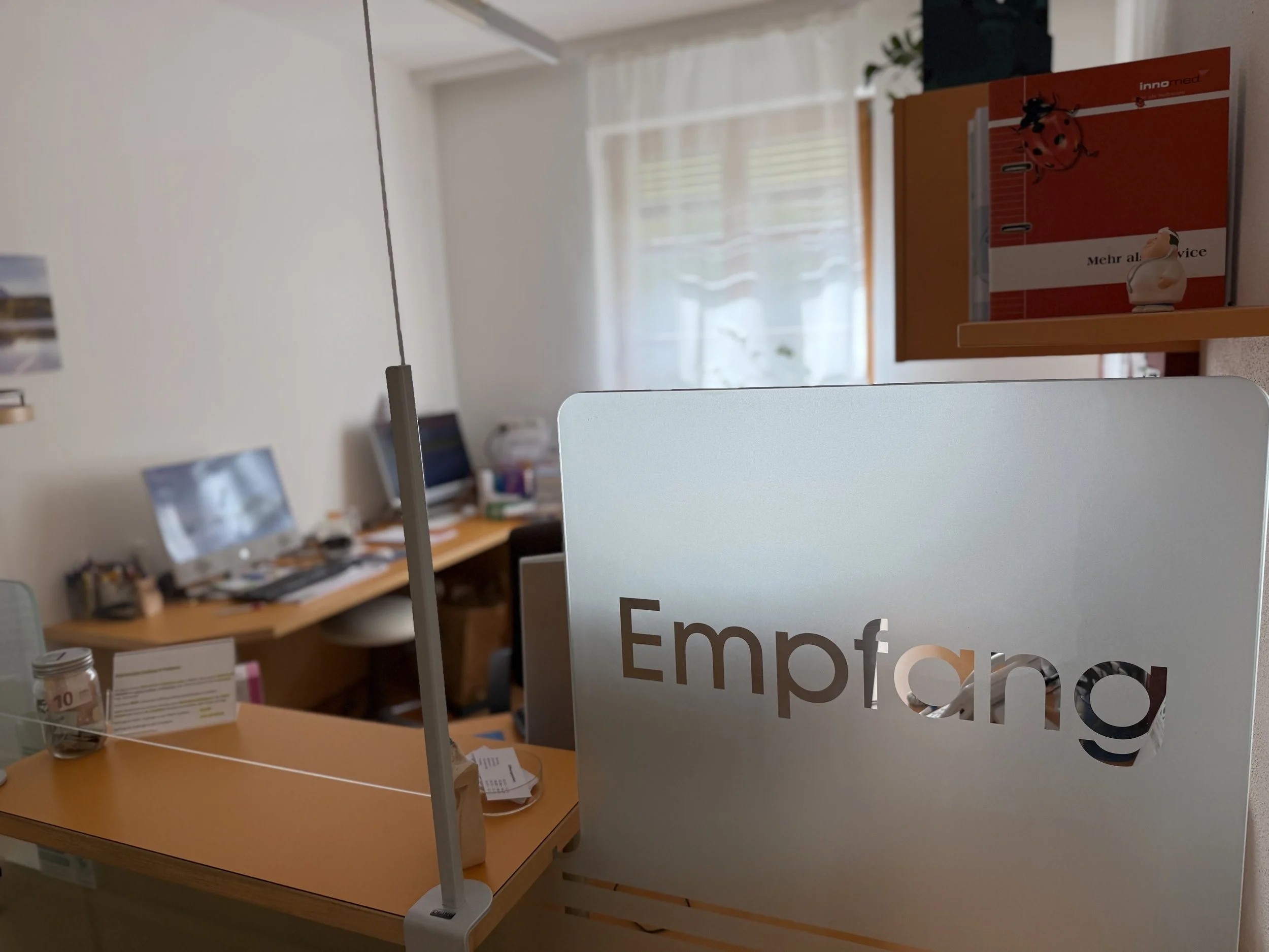 Blick auf einen Schreibtisch mit einer Trennwand mit ausgeschnittenem Schriftzug ‚Empfang‘, verschiedenen Unterlagen, einem Glas und einem Computerbildschirm. Im Hintergrund weitere Computer, Materialien und ein Fenster mit transparenten Vorhängen