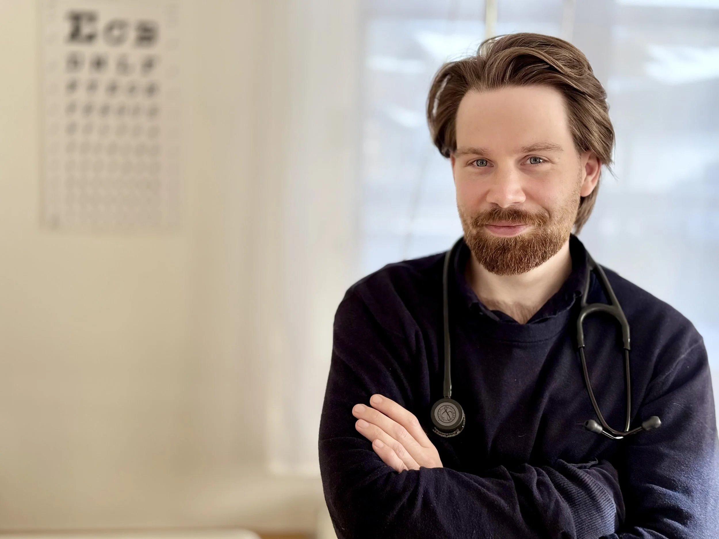 Ein männlicher Arzt mit Bart, der einen dunklen Pullover trägt und ein Stethoskop um den Hals hat, steht mit verschränkten Armen in einem Behandlungszimmer und lächelt in die Kamera.