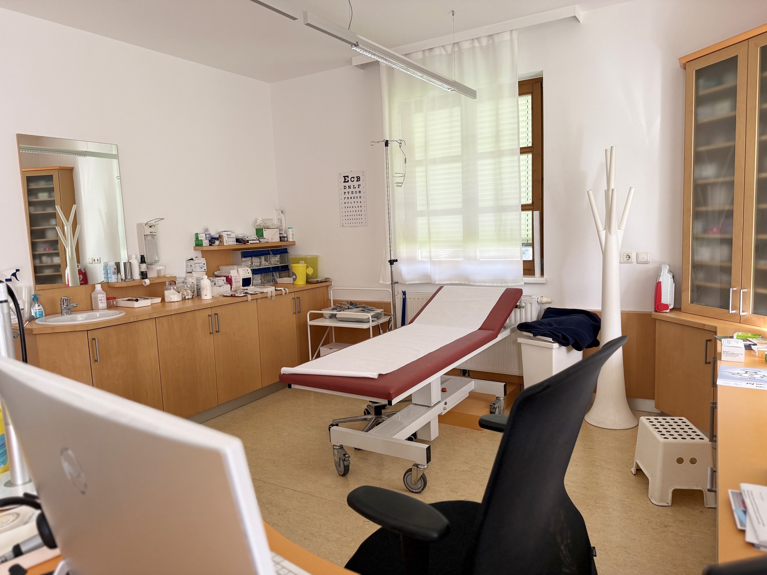 Ein Untersuchungsraum der Ordination mit einer medizinischen Liege, Holzschränken, einem Regal für medizinisches Material, einem Stuhl und einem Fenster mit weißen Vorhängen