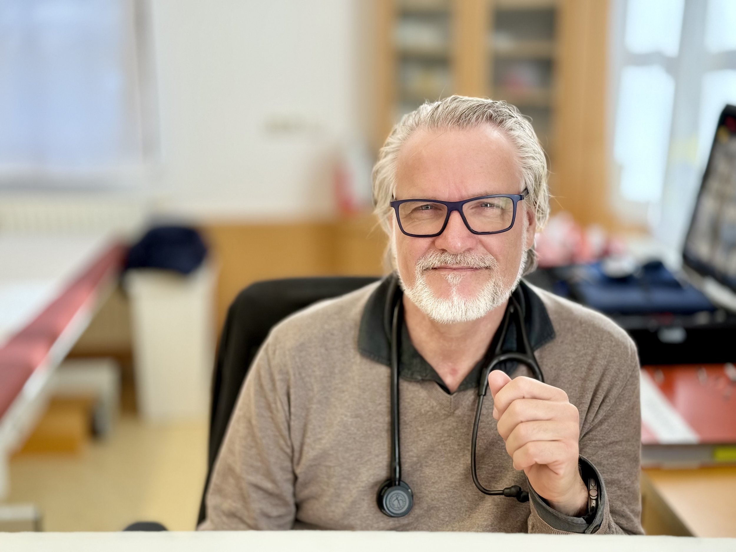 Ein älterer Mann mit grauen Haaren, Bart und Brille sitzt an einem Schreibtisch in einem gut beleuchteten Büro und trägt ein Stethoskop um den Hals.