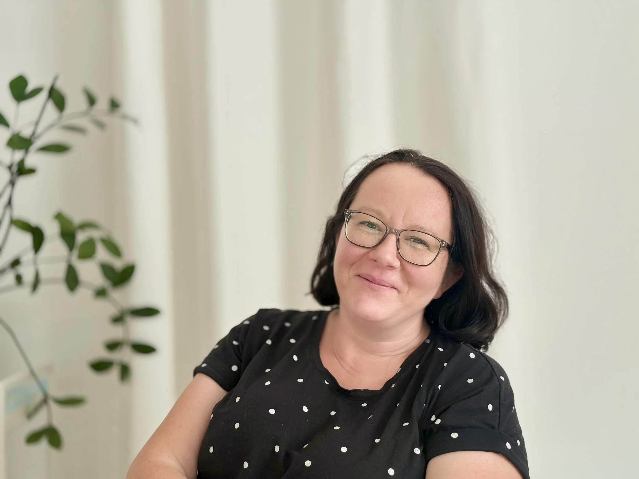 Eine Frau mit schwarzen Haaren, Brille und einem schwarzen Polka-Dot-Shirt lächelt in die Kamera. Sie sitzt in einem Innenraum neben einem weißen Vorhang und einer grünen Zimmerpflanze
