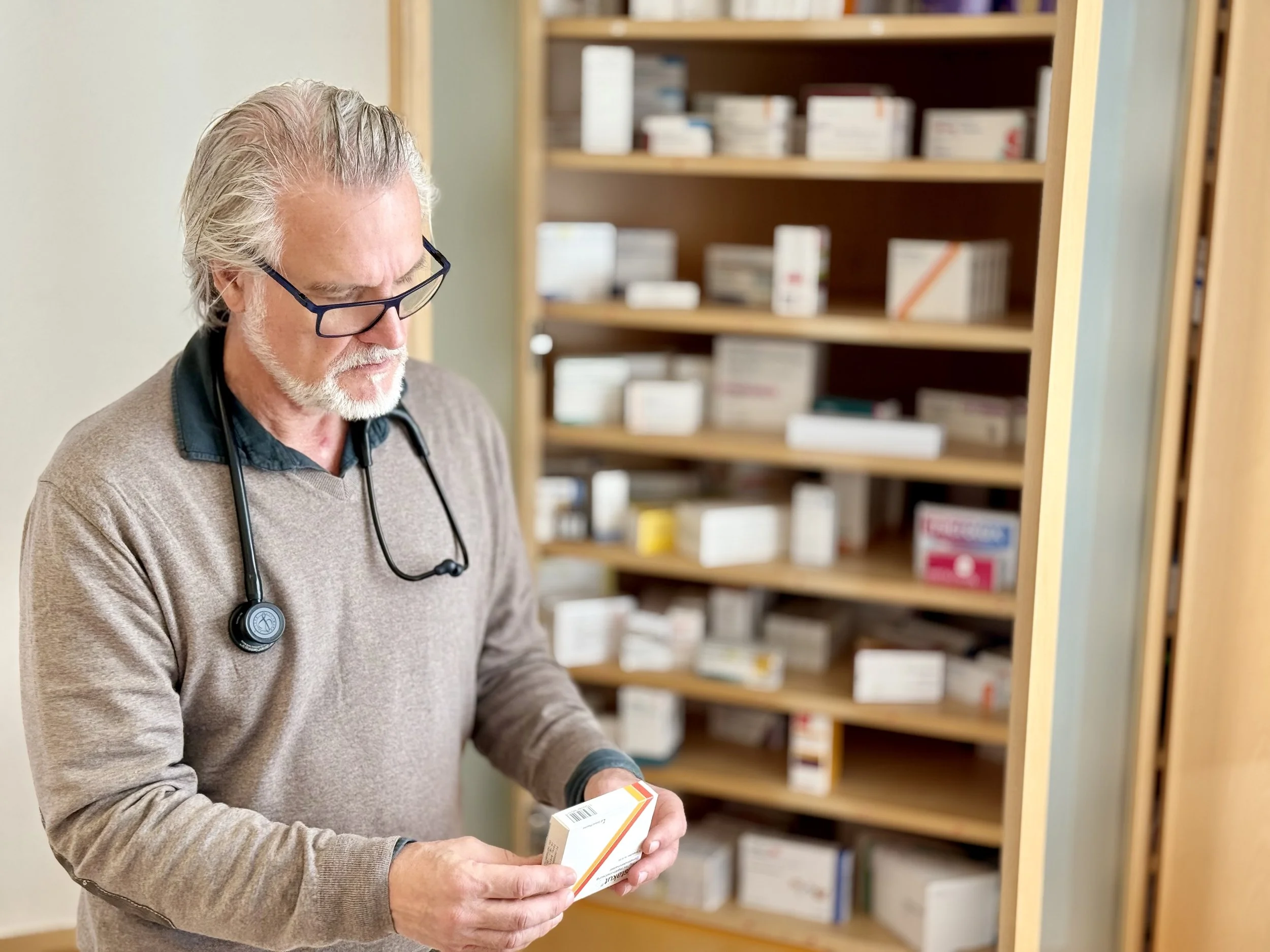 Ein älterer Mann mit grauen Haaren, Brille und Bart, der einen Pullover trägt und ein Stethoskop um den Hals hat, untersucht in einer Apotheke eine Medikamentenpackung. Hinter ihm befinden sich Regale mit Arzneimitteln.