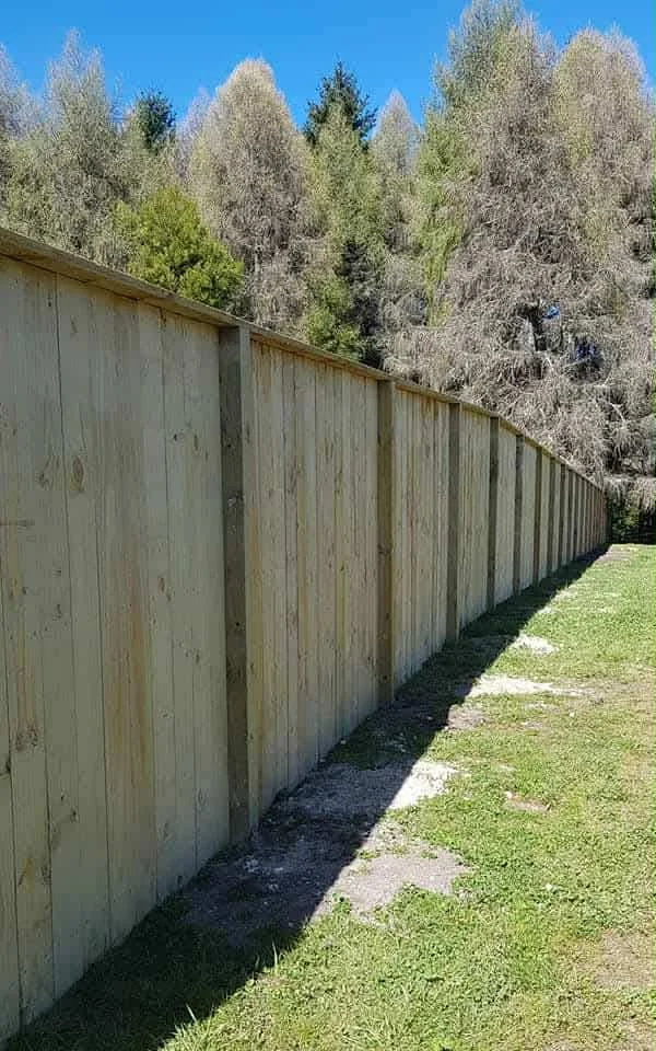 A long wooden privacy fence running along a grassy area with trees in the background under a clear blue sky.