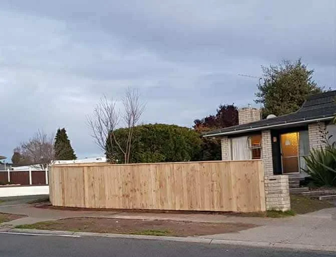 A wooden fence surrounding a residential house with a window and a garden in front, overcast sky.