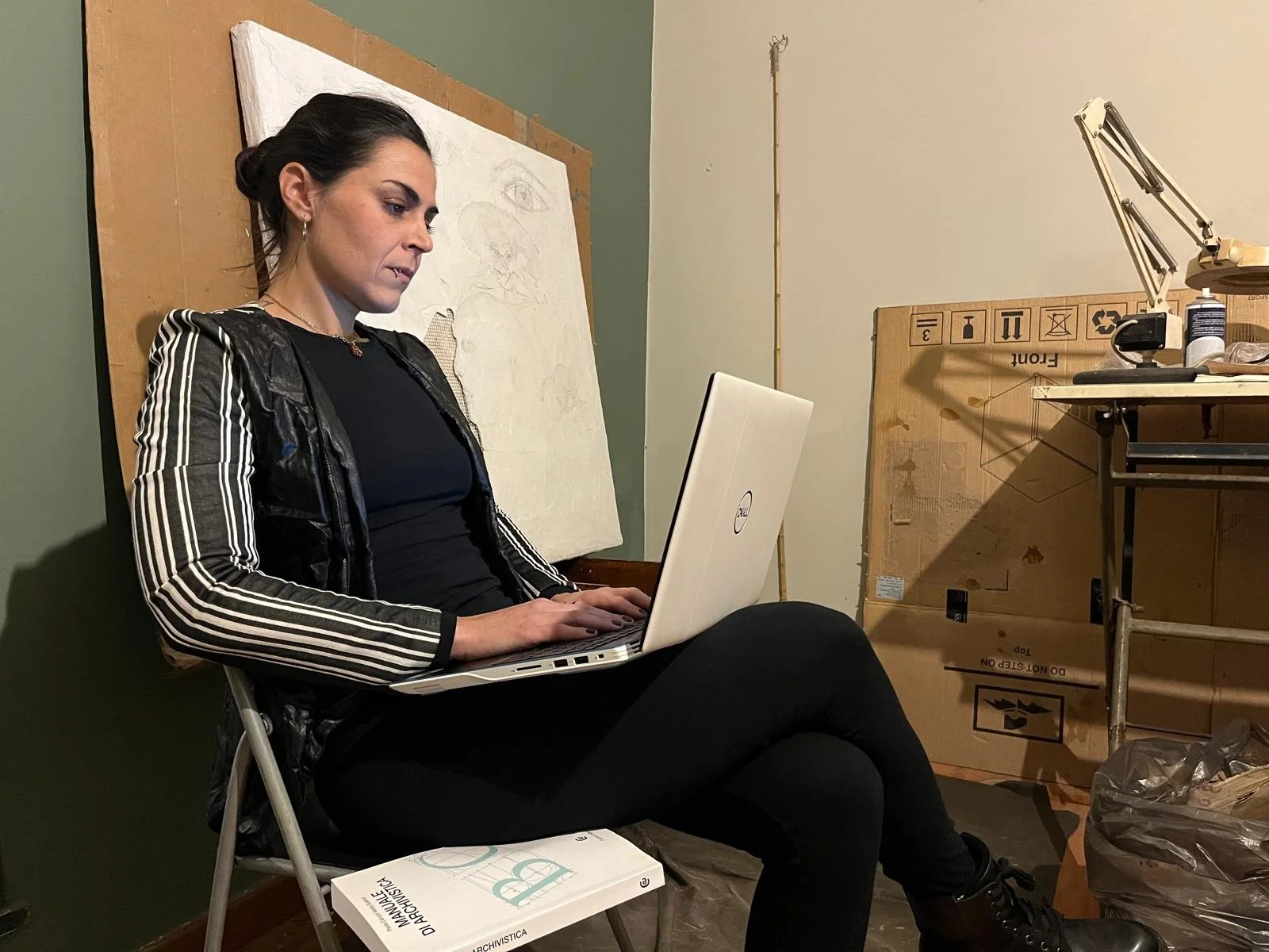 Una donna seduta su una sedia che lavora su un laptop, in un ambiente con materiali artistici e scatole di cartone.