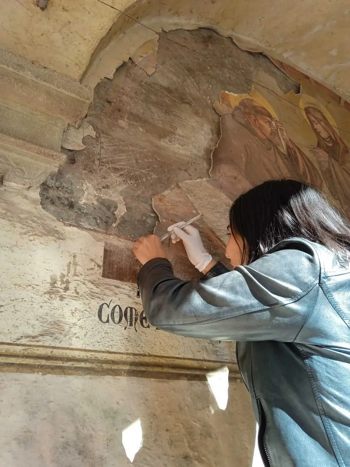 Una donna che lavora su un affresco antico, restaurandolo con pennelli e strumenti, all'interno di un edificio storico.