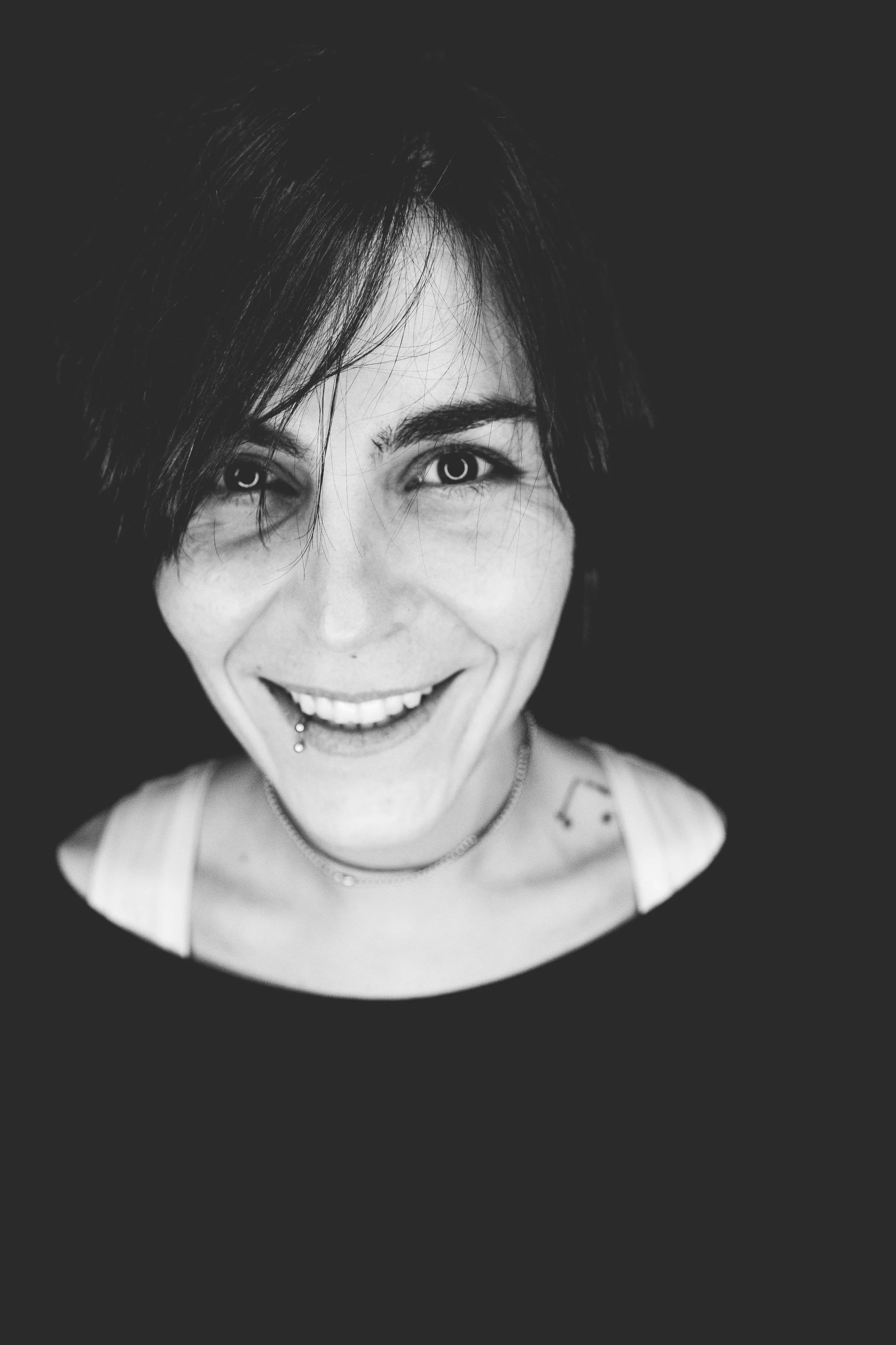 Ritratto in bianco e nero di una donna con capelli scuri, sorriso, piercing alla lingua, collana e tatuaggio sulla spalla, sfondo nero.