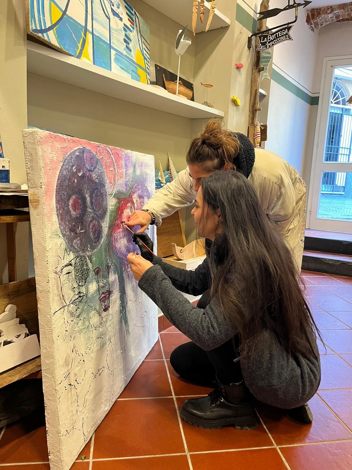 Due donne stanno lavorando insieme su un dipinto astratto su una tela bianca, con colori vivaci e linee disordinate, in un ambiente artistico.