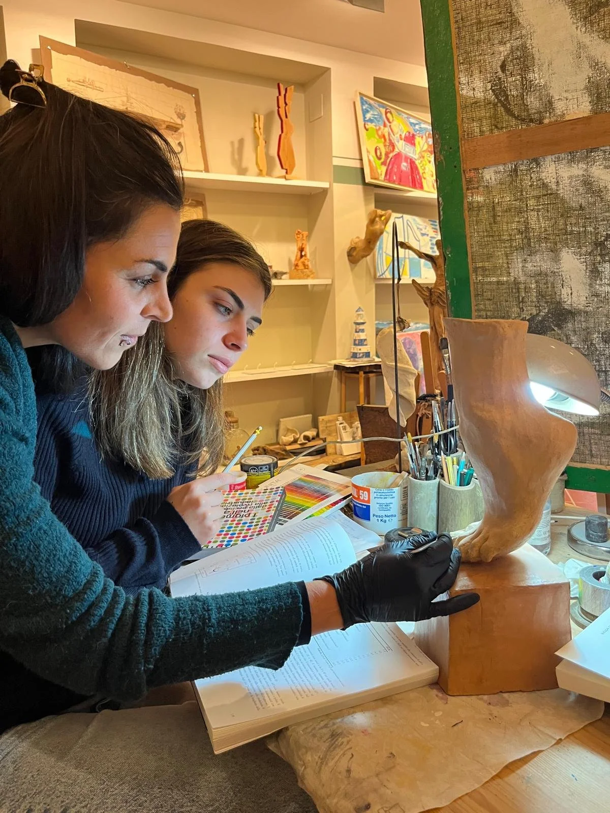 Due donne lavorano su una scultura di legno in un laboratorio artistico, circondate da strumenti e materiali artistici.