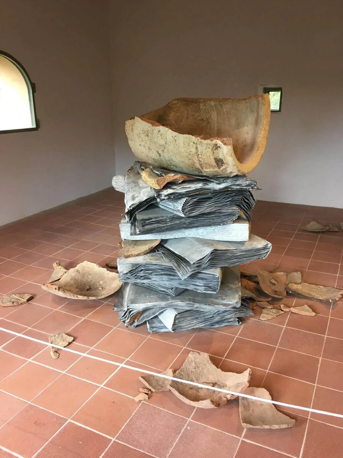 Scultura composta da pagine di giornale sovrapposte e ciotole di terracotta rotte nello spazio di una stanza con pareti beige e pavimento in piastrelle di terracotta.