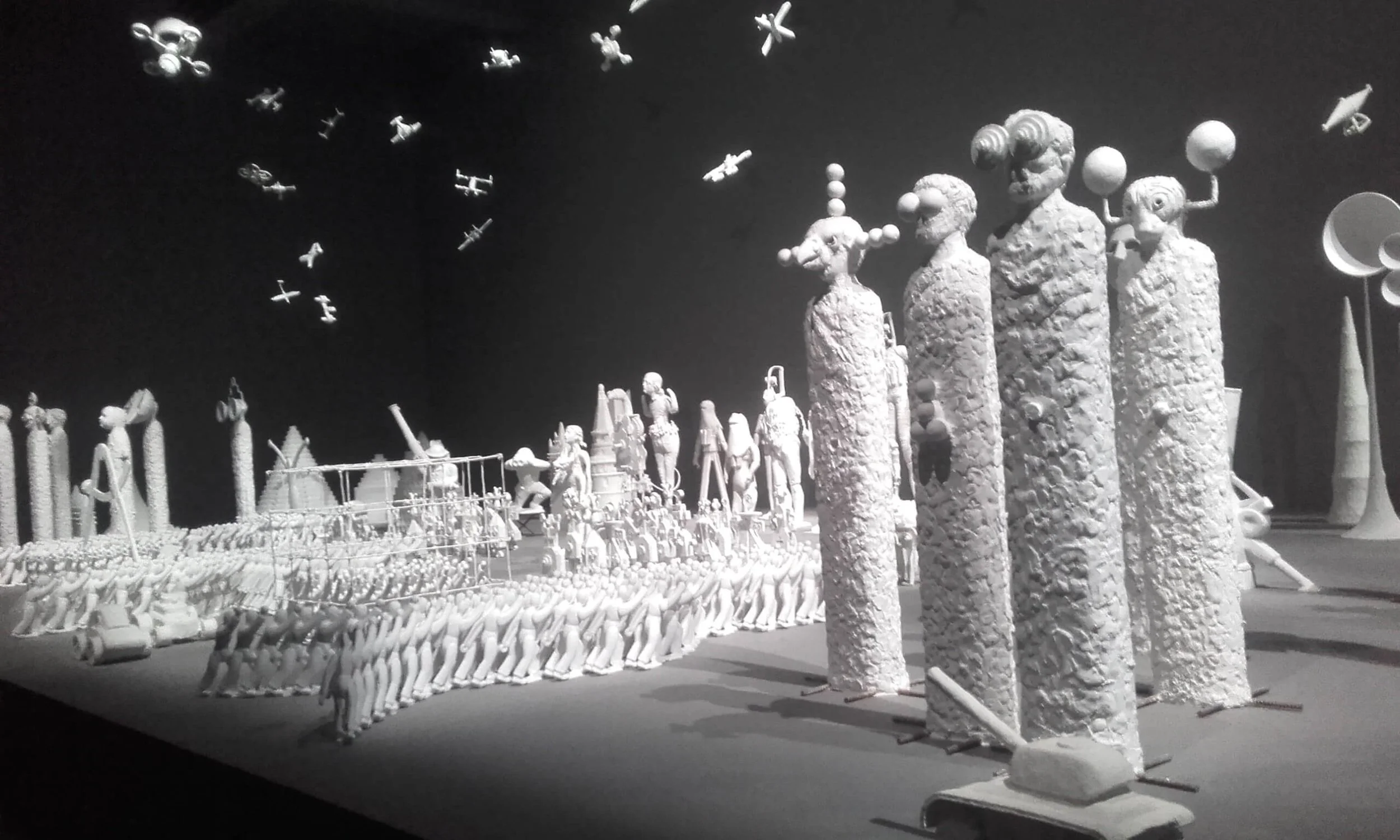 Scultura artistica di figure umane stilizzate e aeroplani in volo, in un ambiente museale in bianco e nero.