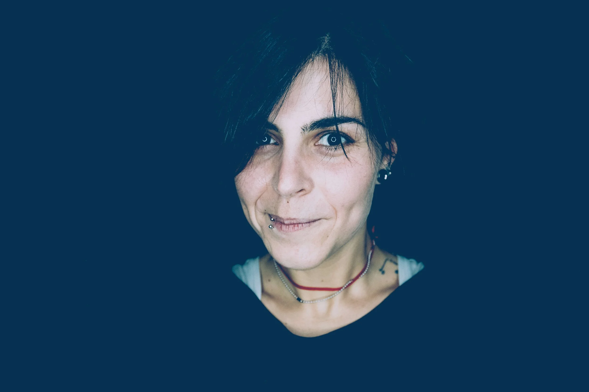 Ragazza con capelli neri, sorriso e piercing alle labbra, indossa collane sottili e orecchini, sfondo nero.