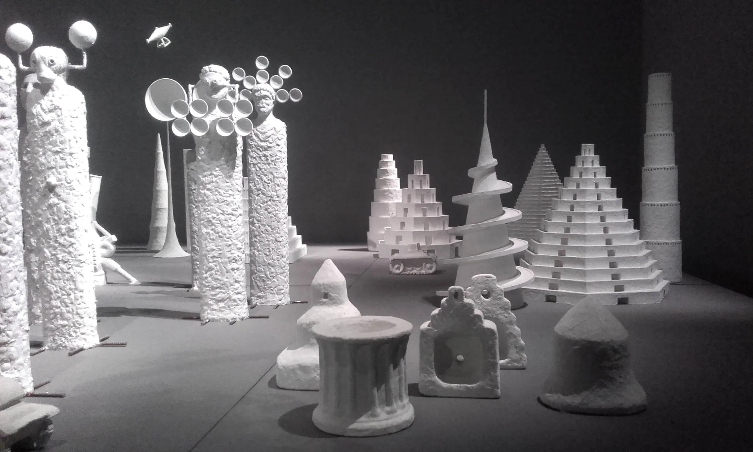 Modellini di oggetti scultorei e strutture futuristiche in bianco in una mostra, con sfondo nero.