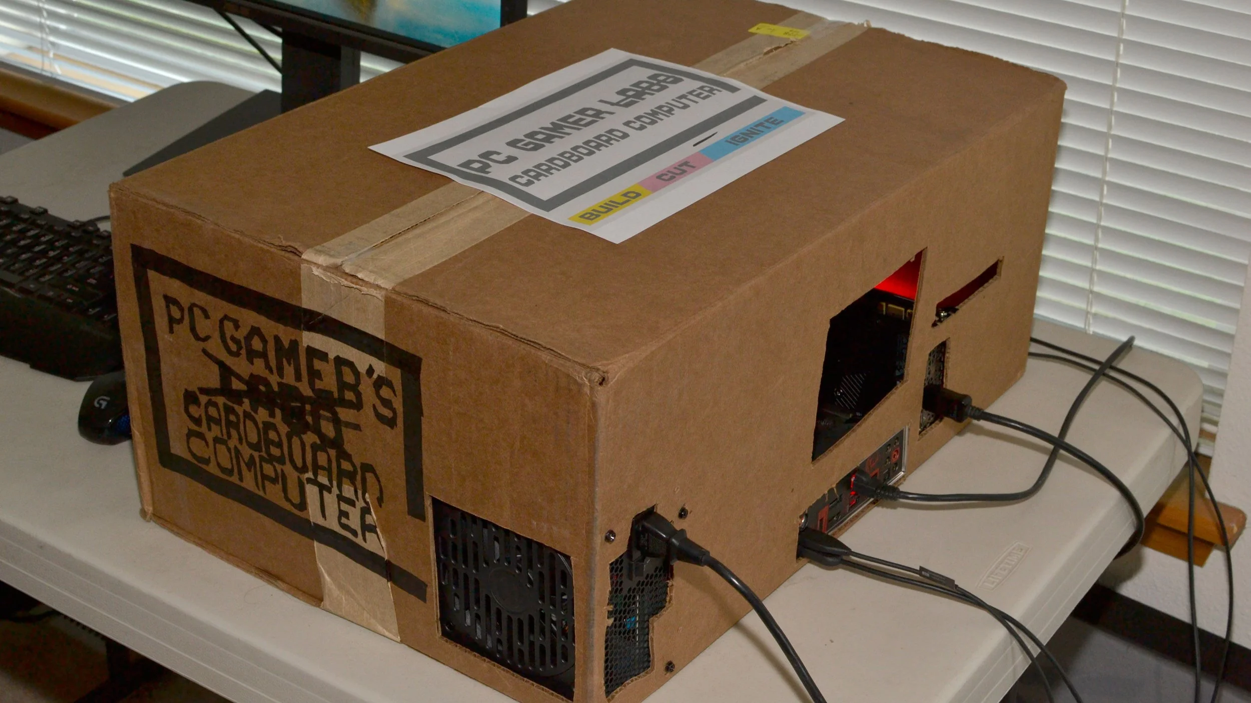 A cardboard box labeled 'PC Gamer's Cardboard Computer' with cutouts on the side revealing a gaming computer inside, connected to power and other cables, placed on a table near a window with blinds.