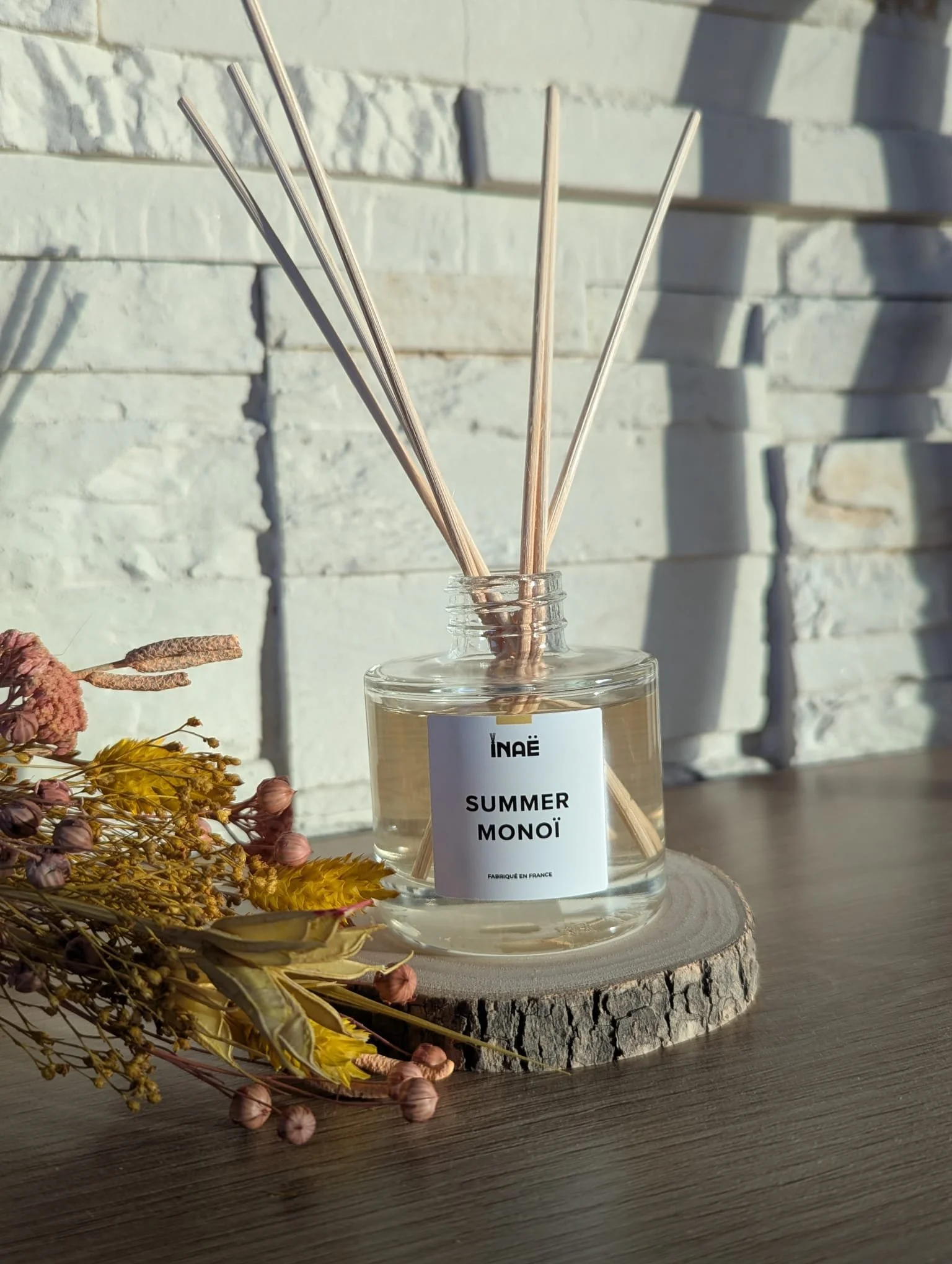 Un diffuseur de parfum avec des bâtons en bois dans un flacon transparent, posé sur une tranche de bois ronde avec des fleurs séchées à côté, contre un mur en briques blanches.
