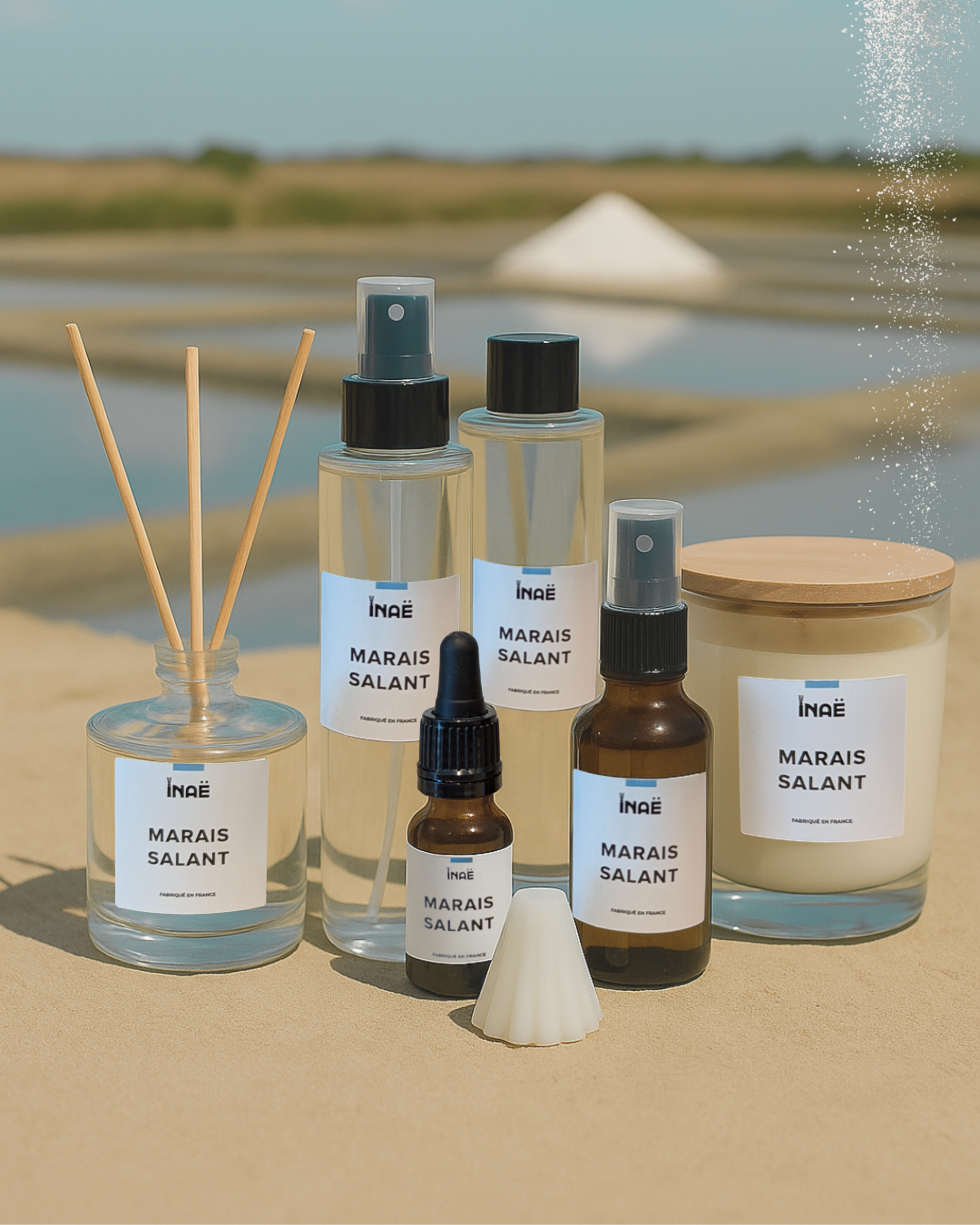 Gamme de produits de soins pour la maison, comprenant des sprays, un diffuseur, une huile, une bougie, un flacon pulvérisateur, un flacon avec pipette, et un râteau de cire parfumée, tous étiquetés 'Marais Salant' sur un fond naturel avec un lac et un saltwork en toile de fond.