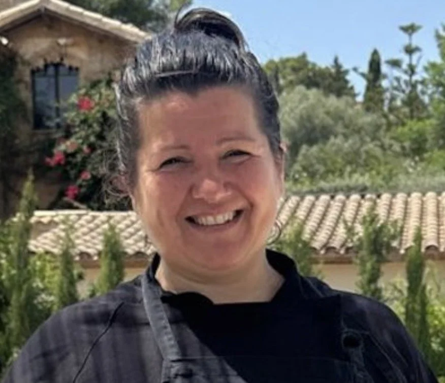 A woman smiling outdoors on sunny day, standing in front of a garden with plants and a house with a tiled roof.