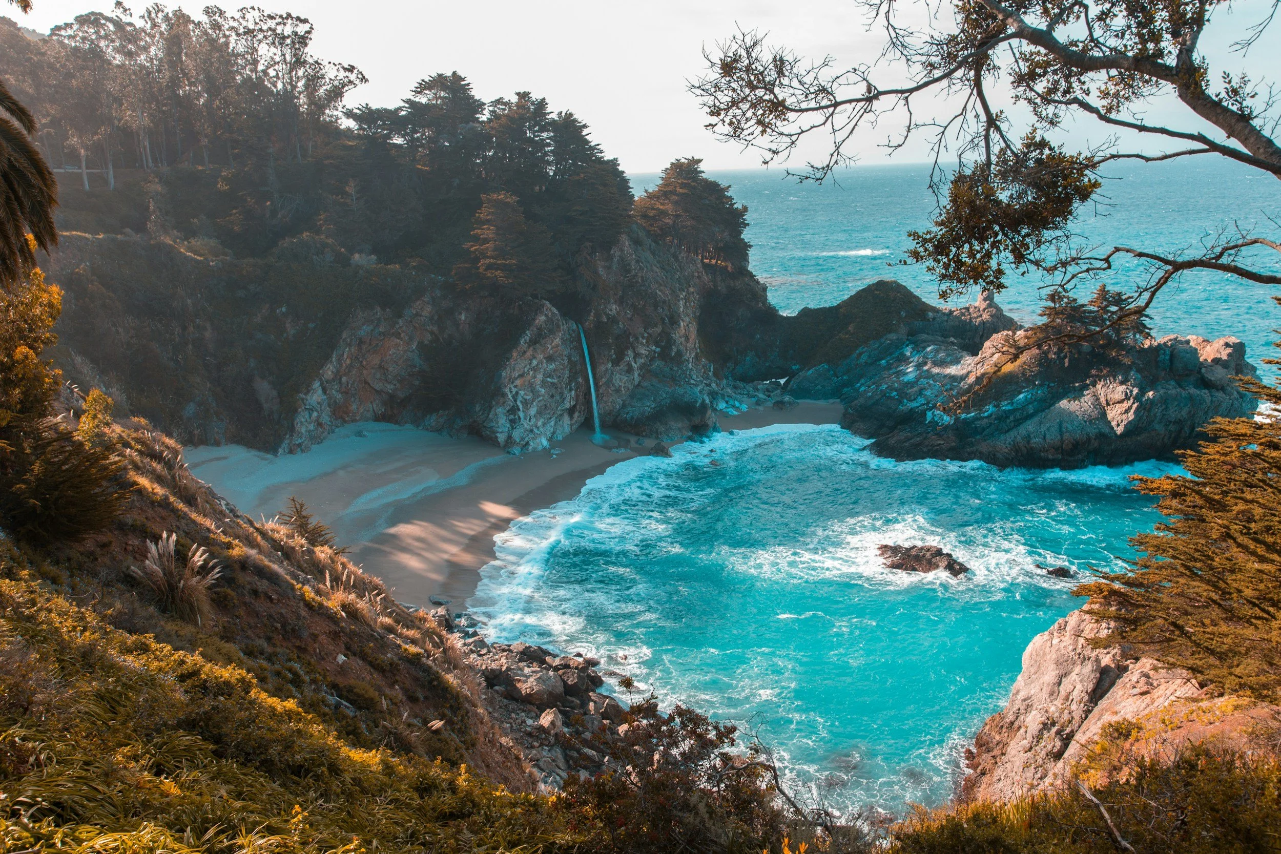 A scenic coastal landscape with a waterfall, rocky cliffs, a sandy beach, and the ocean in the background.