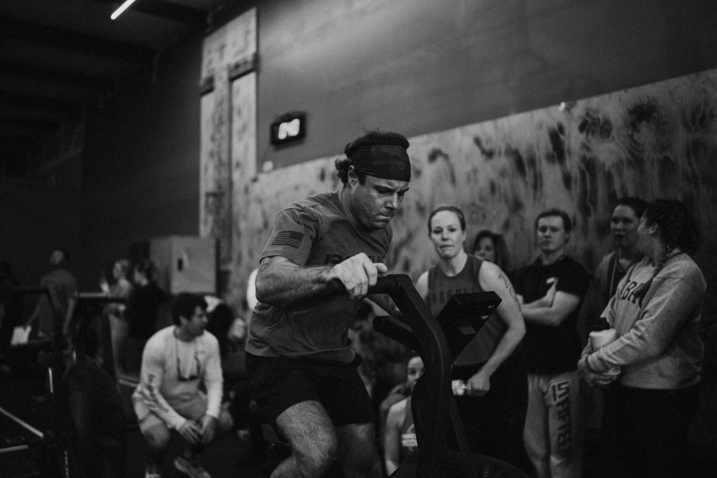 A man with a headband and athletic clothing uses a rower machine while a group of women watches in a gym.