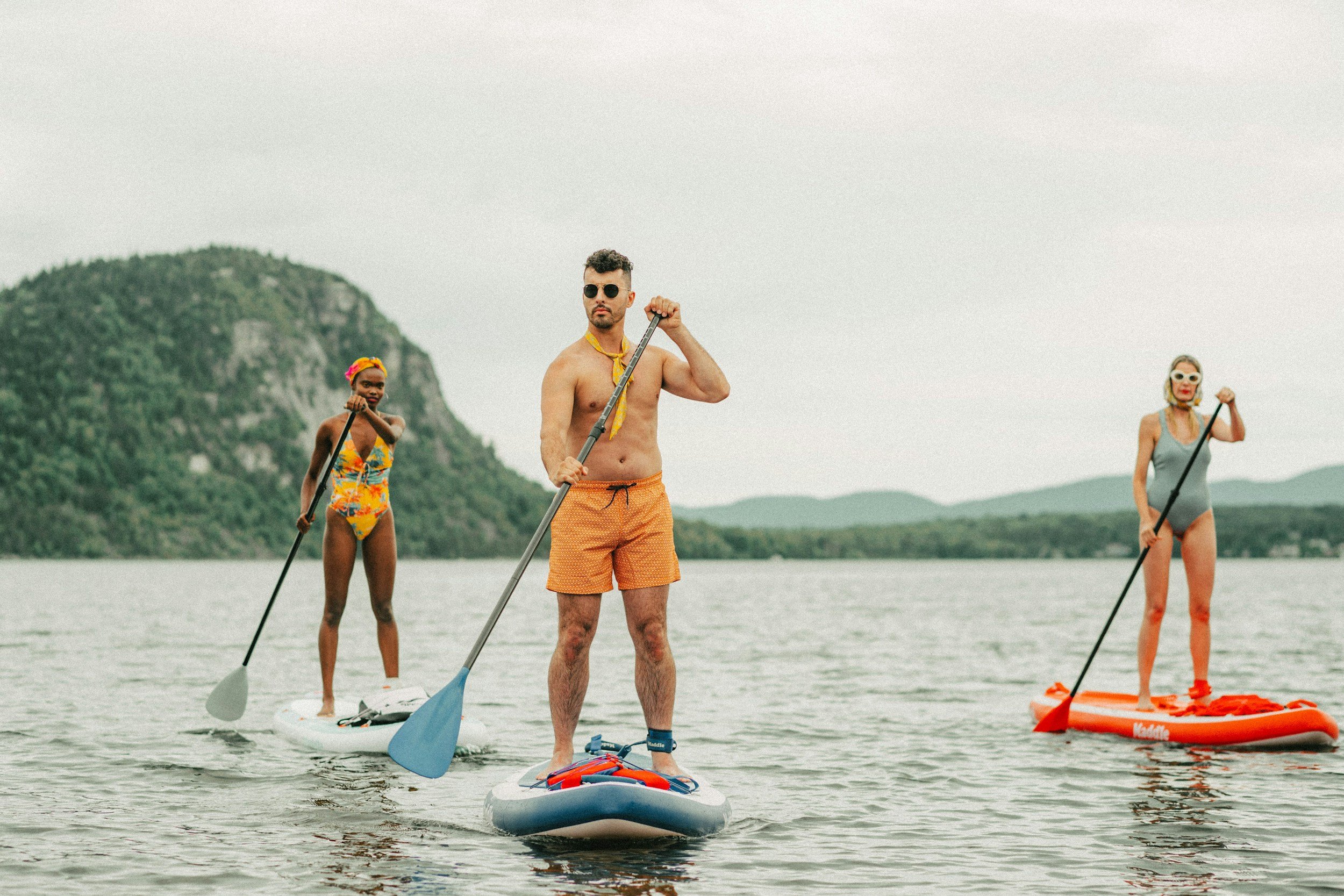 Three people paddleboarding on a lake with a mountain in the background. The man in the center is shirtless wearing orange swim trunks and sunglasses. The woman on the left is in a colorful swimsuit with a headband. The woman on the right is in a gray swimsuit with sunglasses.