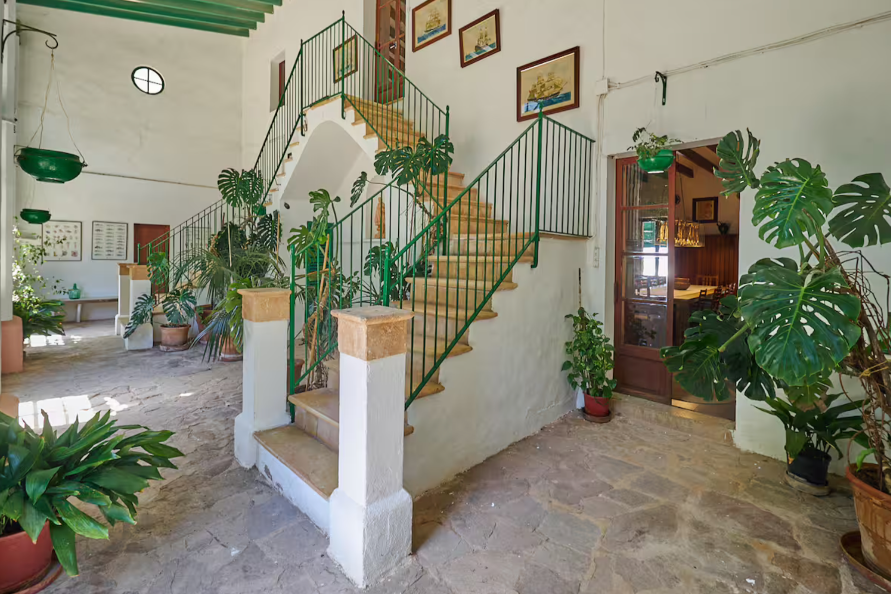 Interior of a bright, rustic house with a stone floor, white walls, and green accents. There are potted plants, a staircase with green metal railings, and framed artwork on the wall. An open doorway reveals a dining area with wooden furniture.