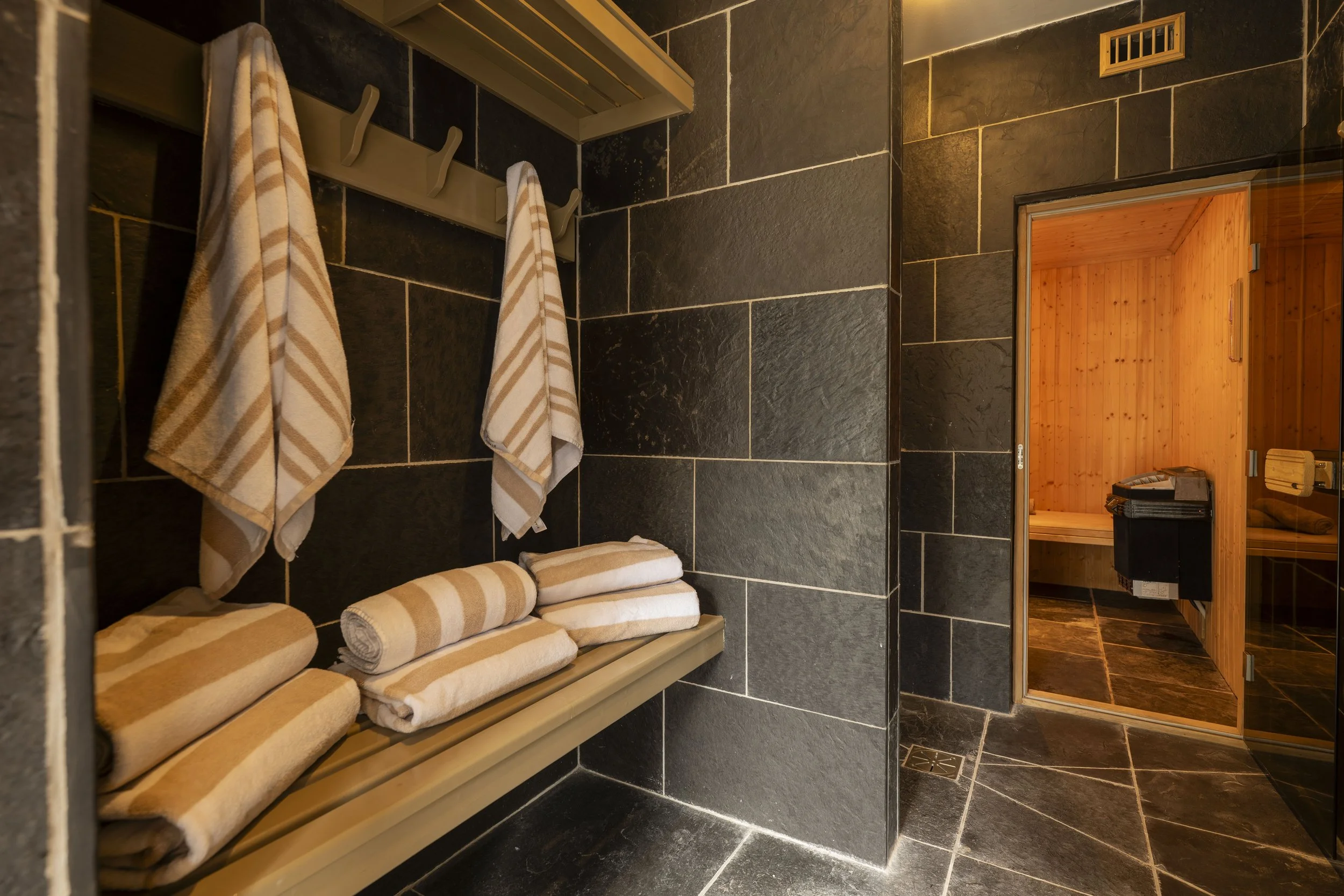 A steam room with black tiled walls and floor, towels and robes hanging on hooks, and a wooden sauna room visible through an open door.