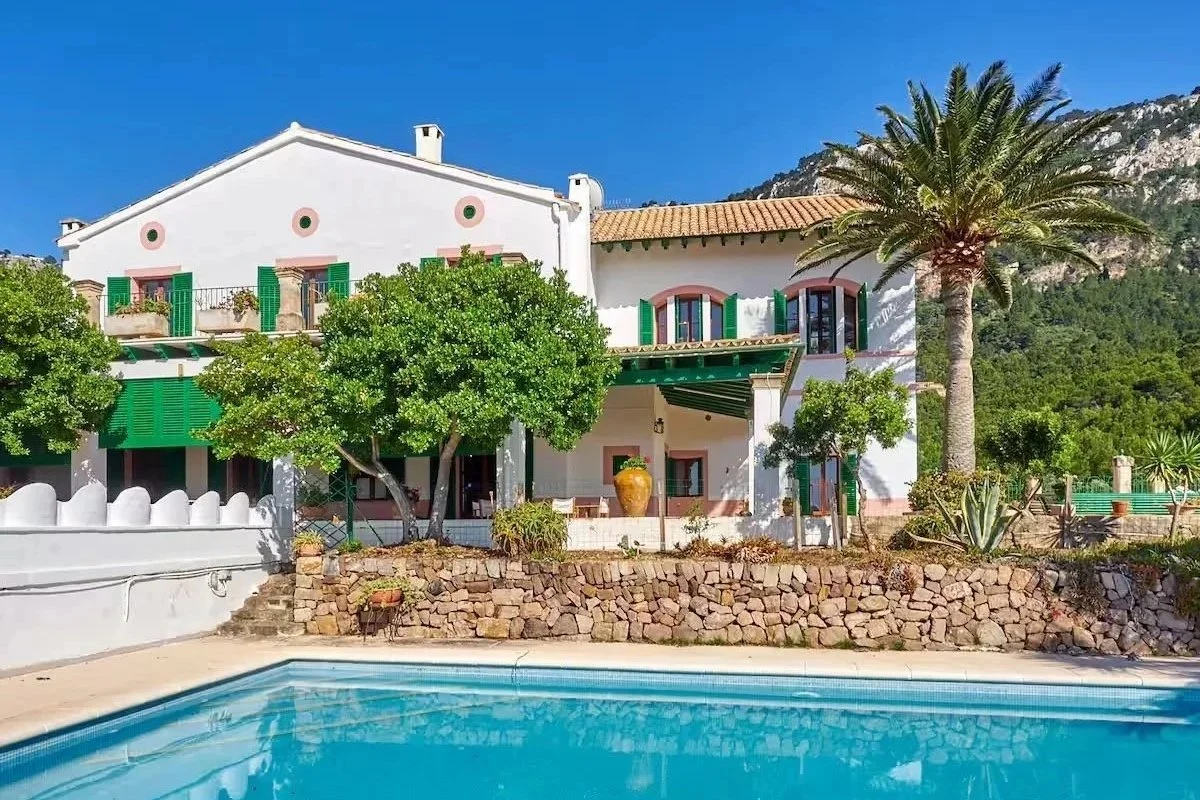 A large white Mediterranean-style house with green shutters, a swimming pool in the foreground, and lush green trees and a mountain backdrop.