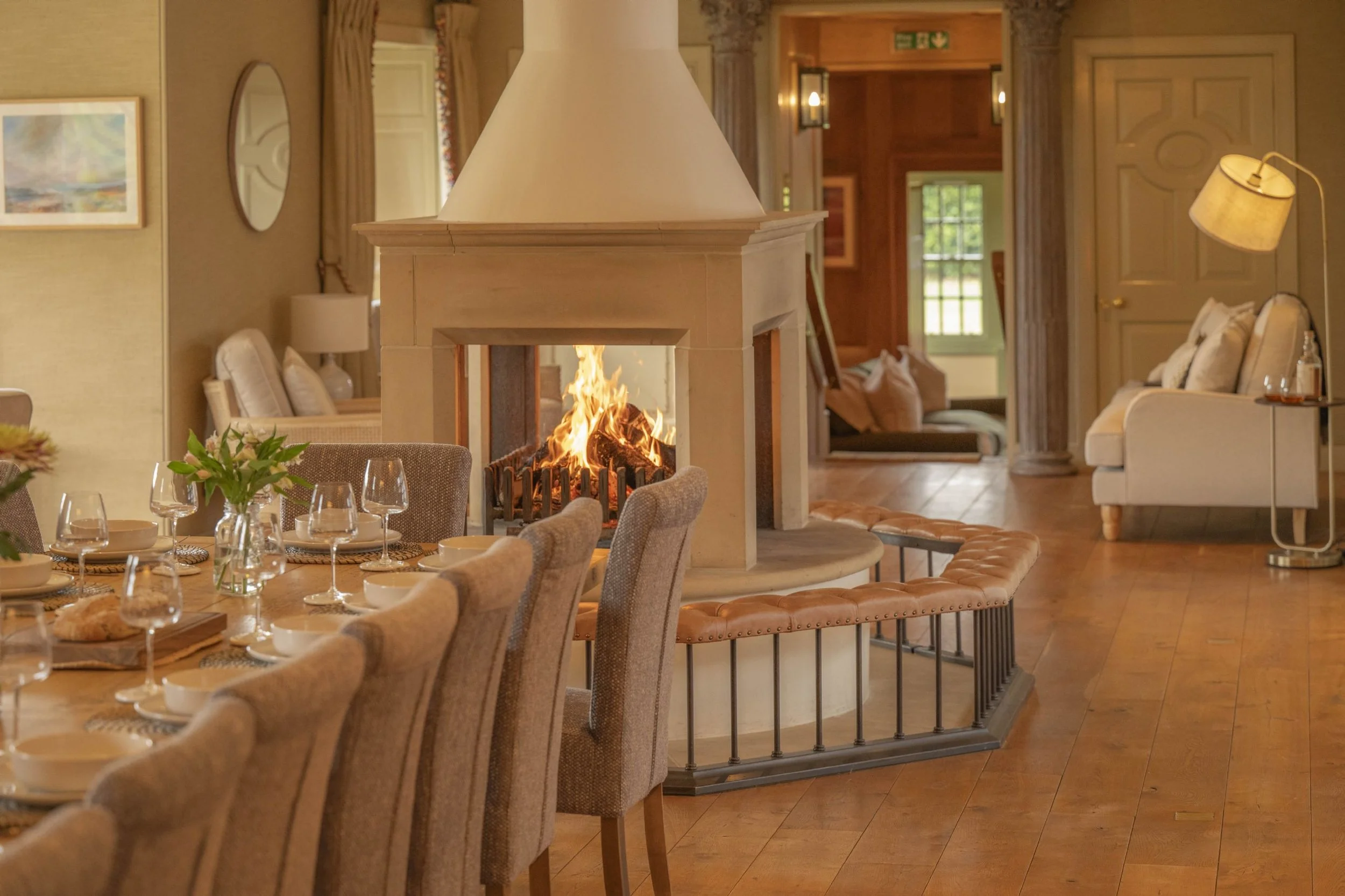 A cozy dining and living room area with a central white fireplace with a fire burning inside. To the left, a dining table is set with plates, wine glasses, and a small vase of flowers. To the right, there is a white sofa, a standing lamp, and some furniture in the background. The room has wooden floors and warm lighting.