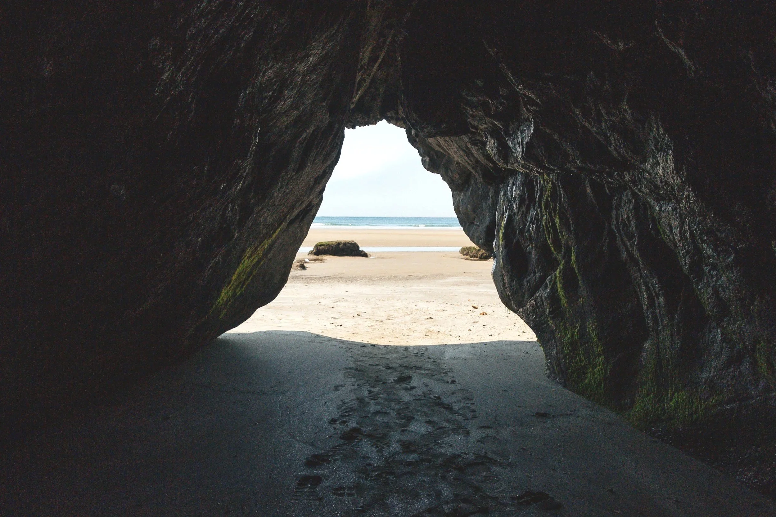 View from inside a rocky cave looking out onto a sandy beach with the ocean in the background.