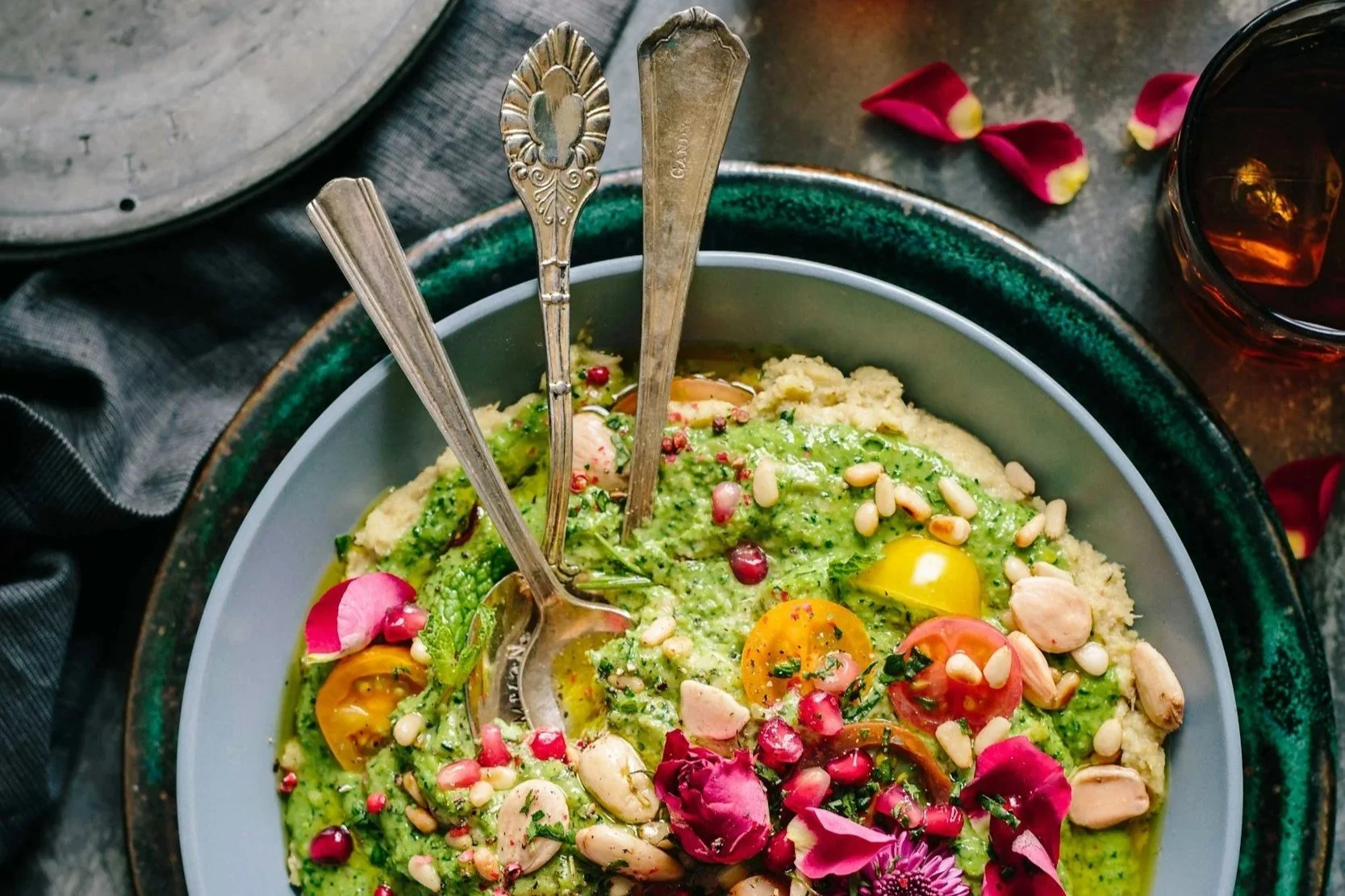 A bowl of green guacamole topped with cherry tomatoes, pomegranate seeds, pine nuts, and flower petals, with three vintage silver spoons inside. There is a glass with a dark beverage to the right and pink flower petals scattered around.