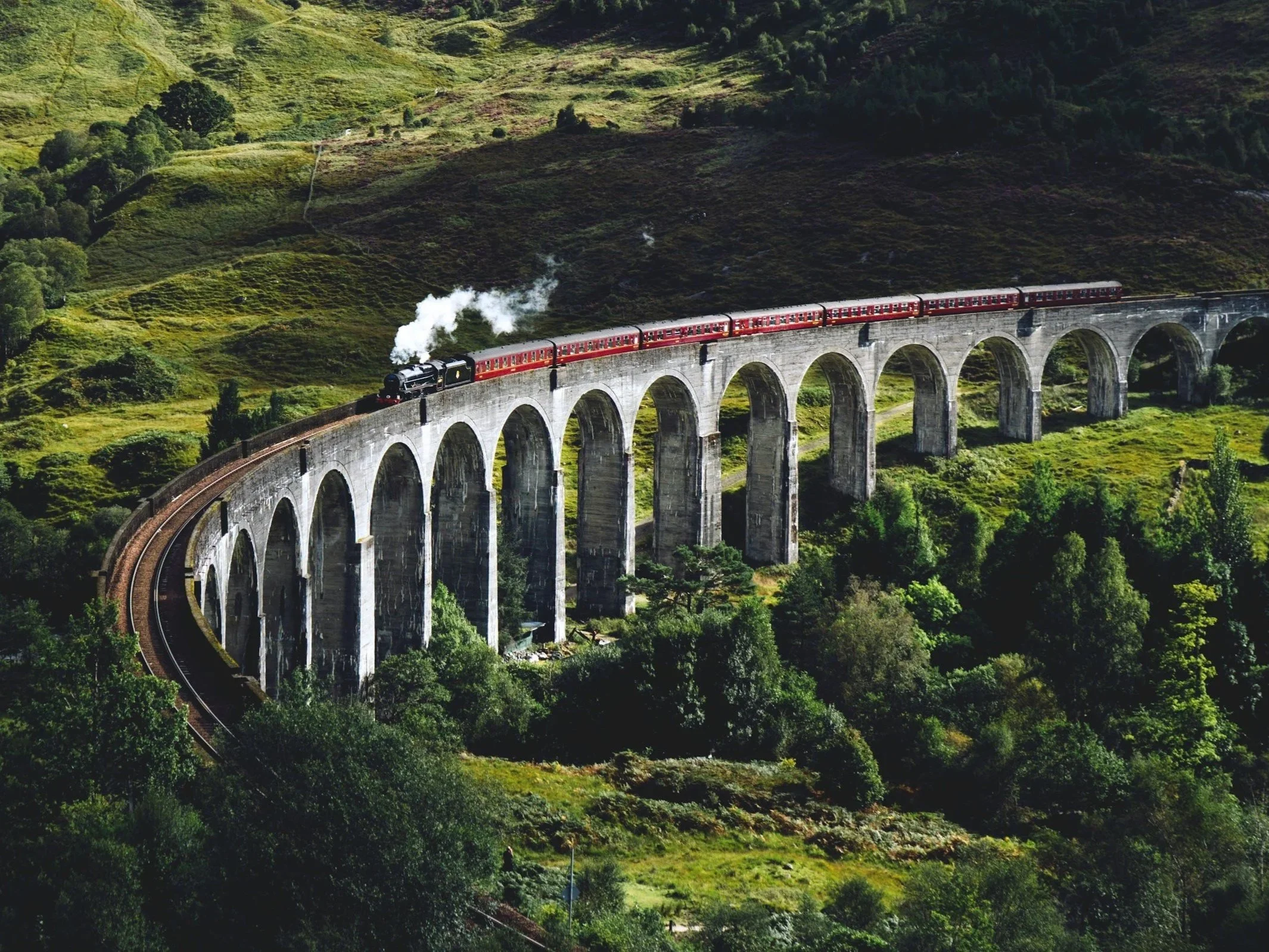 A steam train traveling across a tall stone arched viaduct in a green hills landscape.