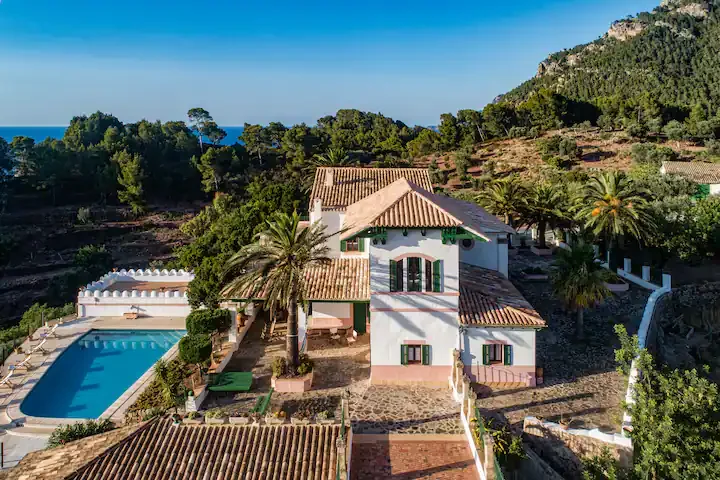 Ville with green shutters, tiled roof, and a covered porch, surrounded by palm trees and greenery, with a swimming pool on the left and hills with trees and the ocean in the background under a clear blue sky.