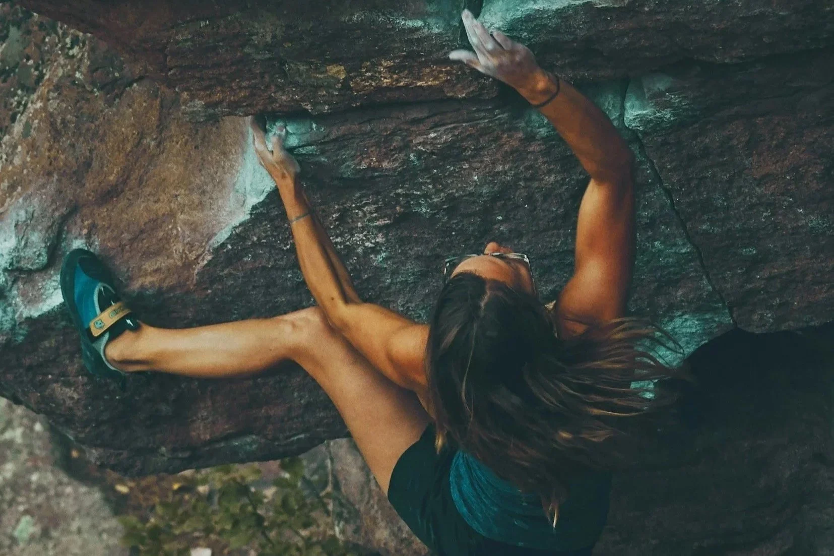 A woman wearing climbing shoes and shorts rock climbing on a vertical rock face with her arms stretched upwards and her head tilted back.