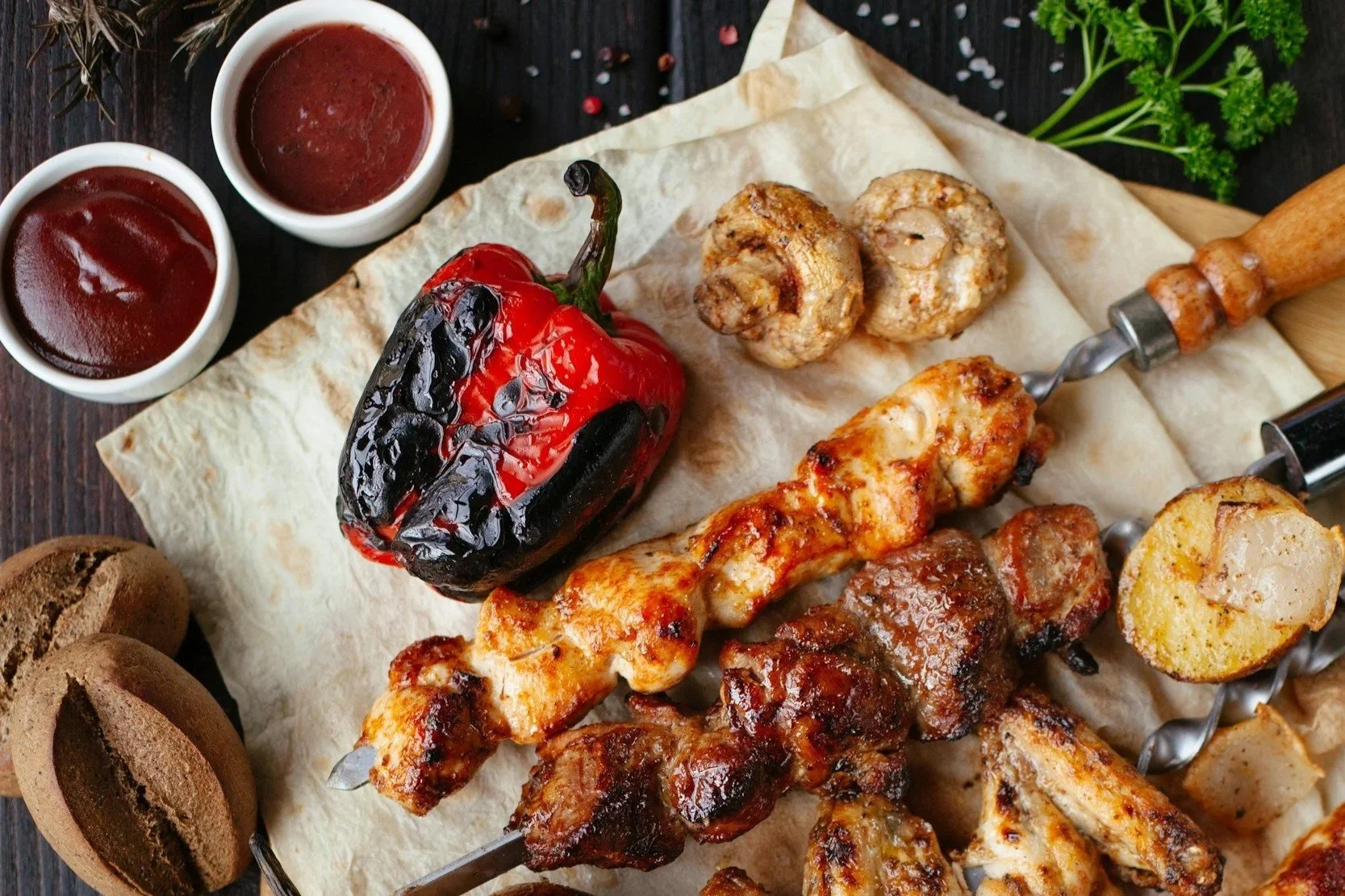 A platter of grilled skewered meats, a grilled red bell pepper, two small bowls of barbecue sauce, two chocolate cookies, and parsley garnish on a wooden table.
